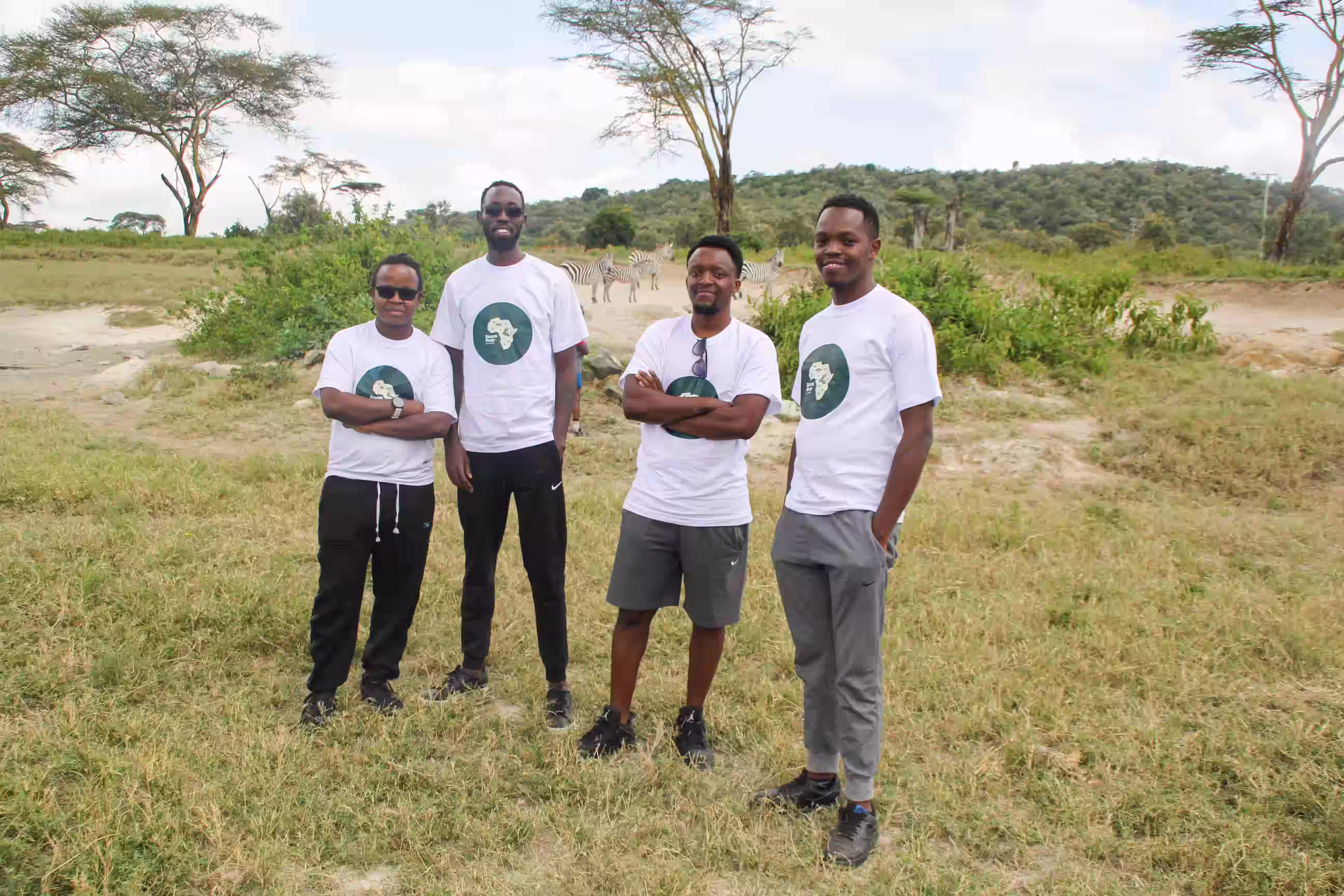 Four men standing on grass in a savanna setting wearing matching roamroar t-shirts, with zebras and trees in the background.
