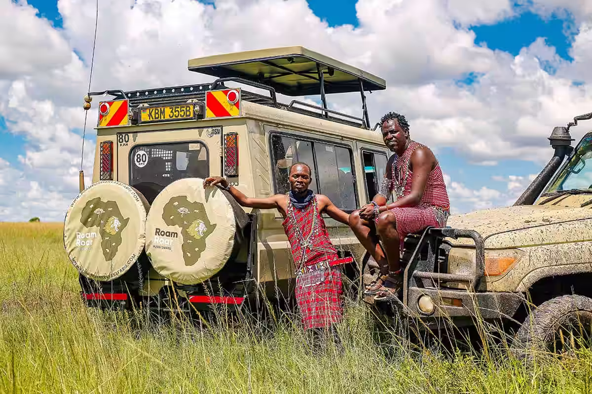 Two men in traditional Maasai clothing posing beside a safari vehicle in tall grass under a partly cloudy sky.
