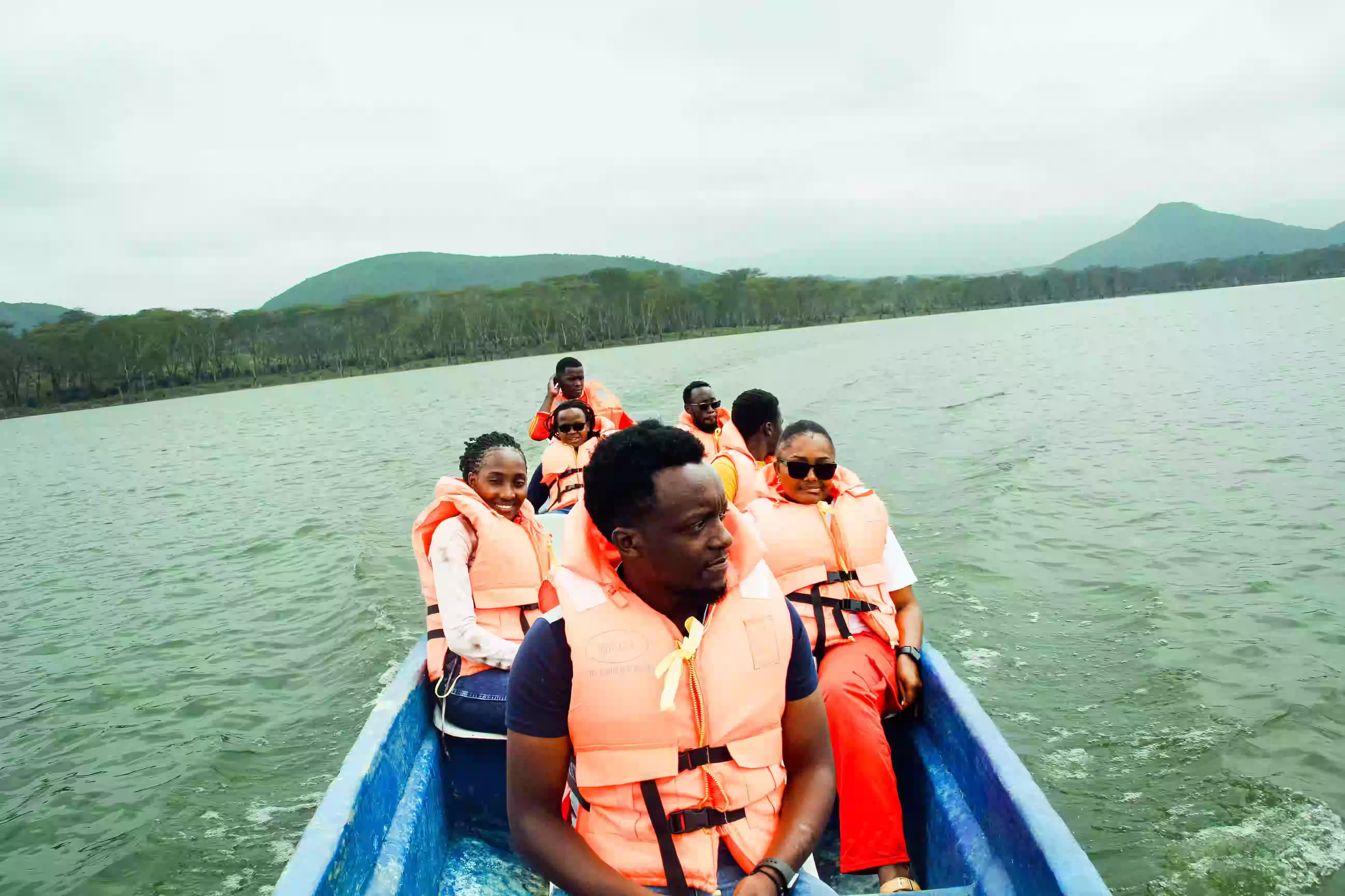 A group of people wearing orange life jackets seated in a blue boat on a lake with forested hills in the background.