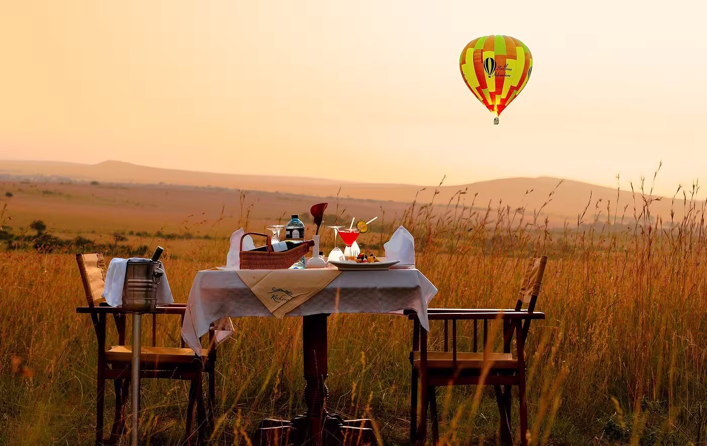 Outdoor table set for two with drinks and snacks in a grassland during sunset, with a colorful hot air balloon floating in the sky.