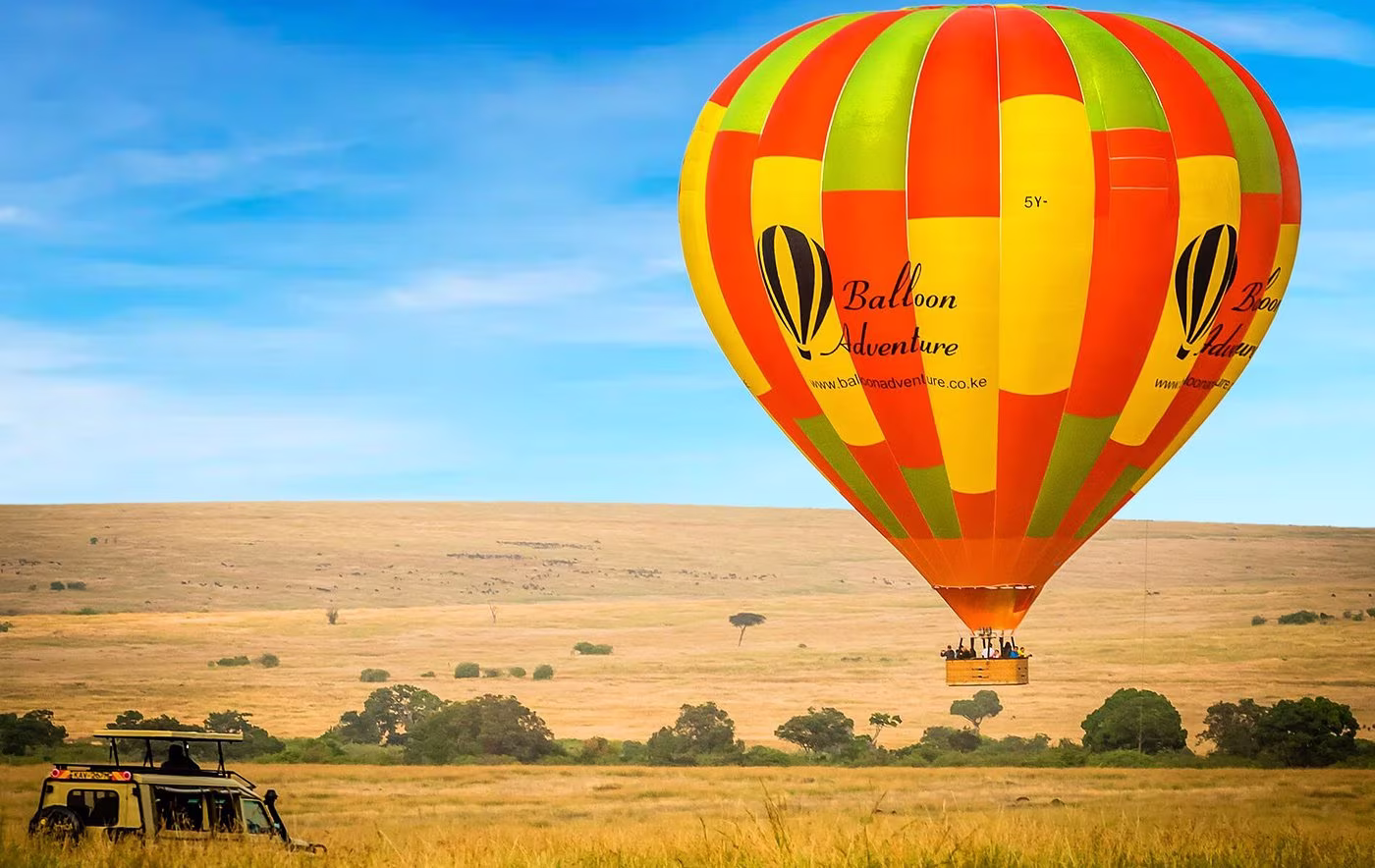 Colorful hot air balloon with passengers flying over a savannah landscape with a safari vehicle below.