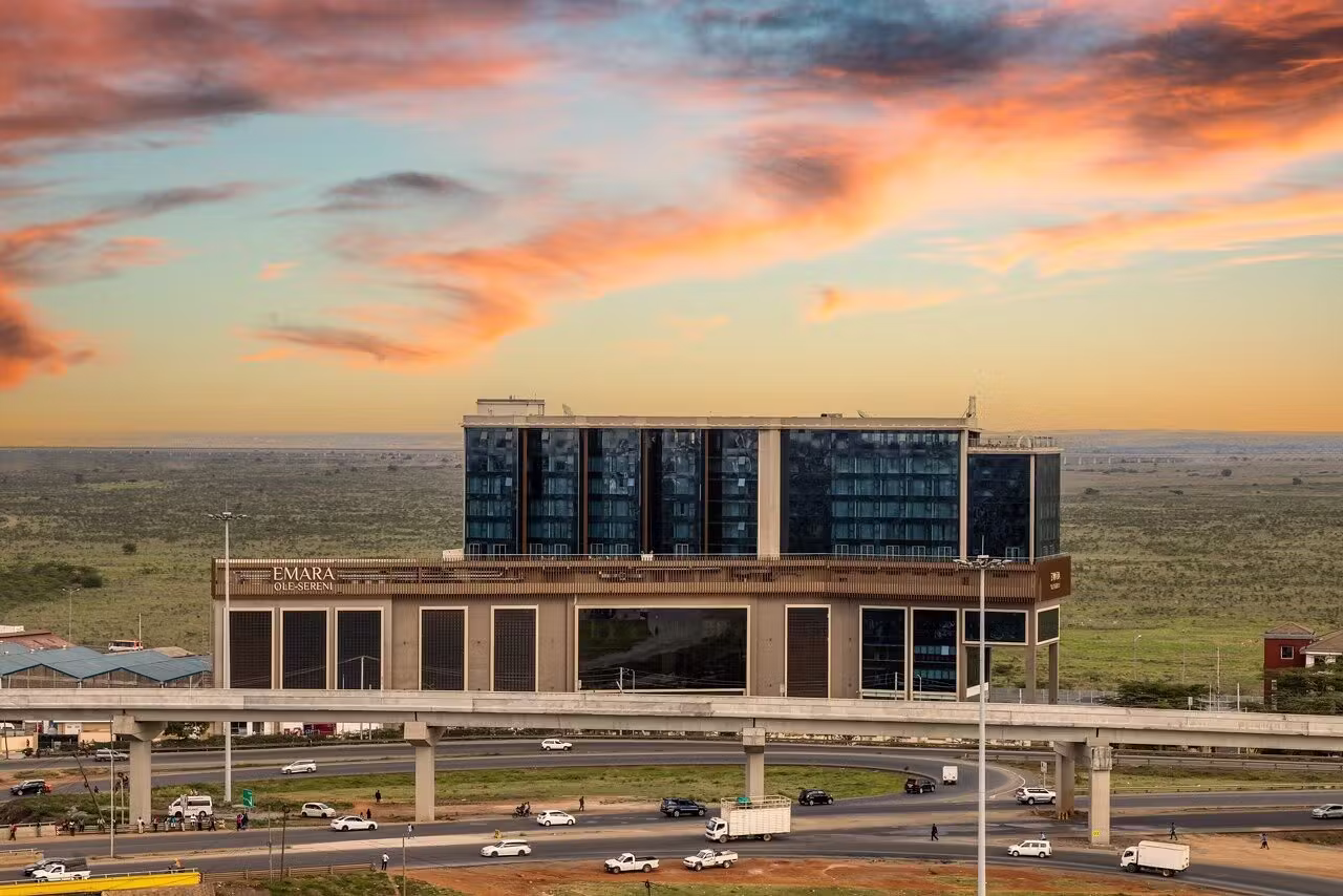 Modern multi-story Emara Ole-Sereni building with glass windows beside highway under a colorful sunset sky.