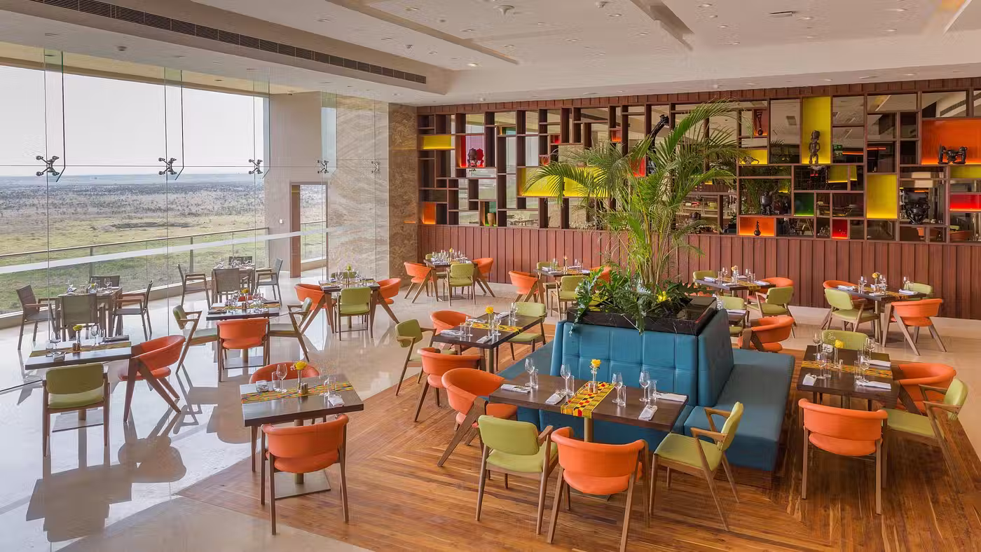 Bright restaurant interior with orange and green chairs, wooden tables, a large central planter with green plants, and floor-to-ceiling windows overlooking a scenic landscape.