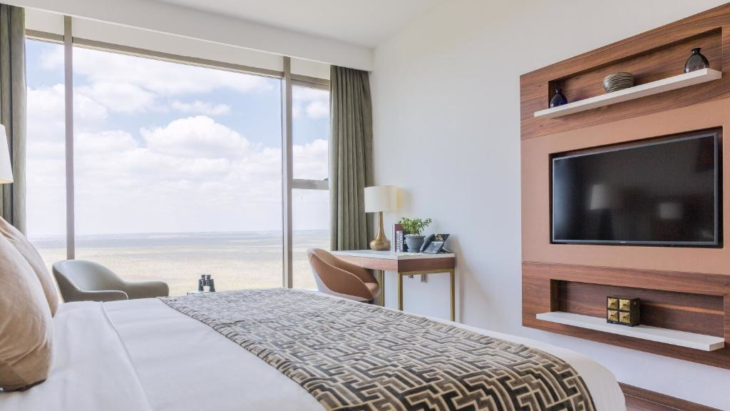 Modern hotel room with a large bed, patterned gray and white bedspread, large window with a view, beige armchairs, desk with lamp and plants, and wall-mounted flat screen TV.