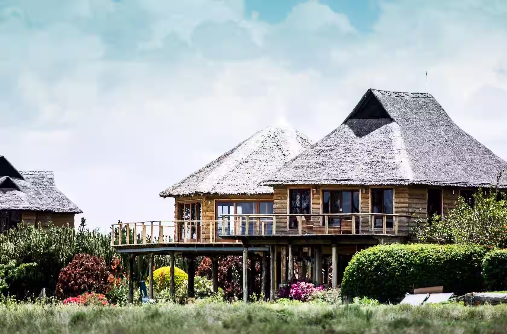 Wooden elevated huts with thatched roofs surrounded by lush green bushes and colorful flowers under a bright sky.