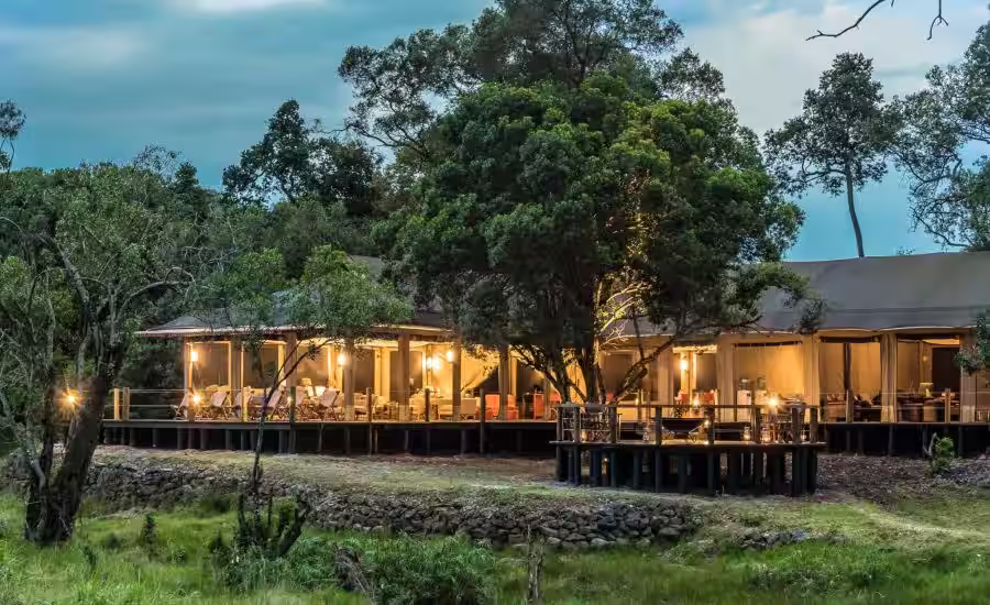 A warmly lit lodge with outdoor seating surrounded by trees and greenery at dusk.