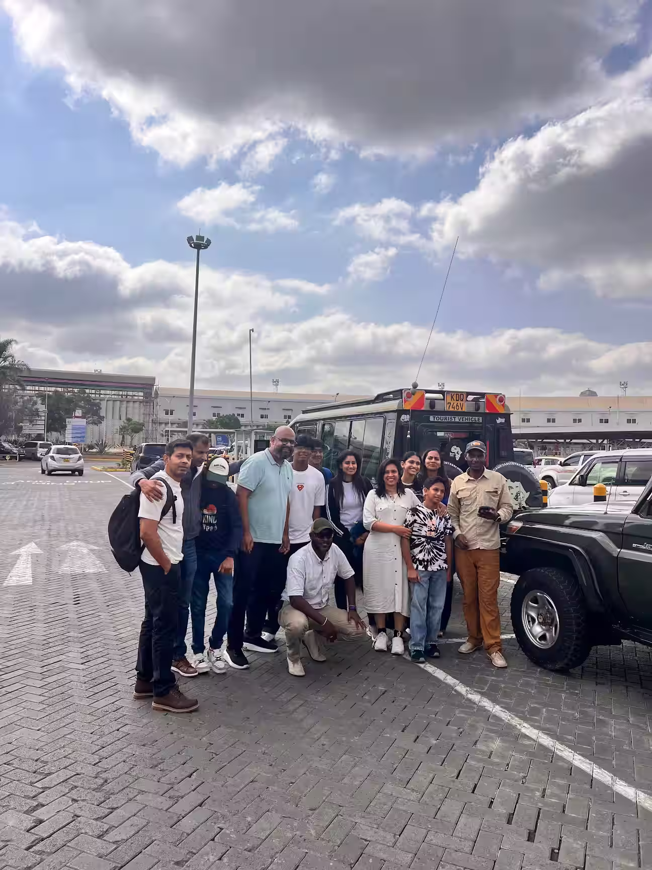 Group of eleven people posing together in front of a tourist vehicle parked in a paved lot under a cloudy sky.