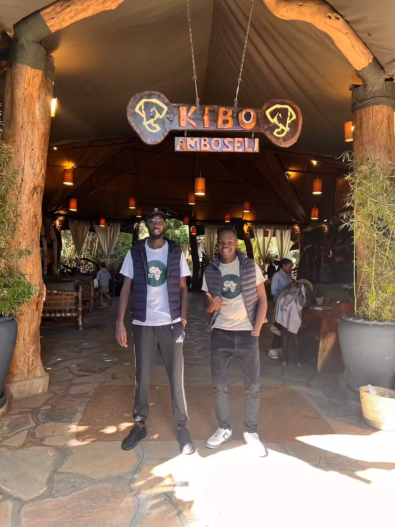 Two young men wearing matching t-shirts and vests standing under a wooden archway with a sign reading 'Kibo Amboseli' at a lodge entrance.