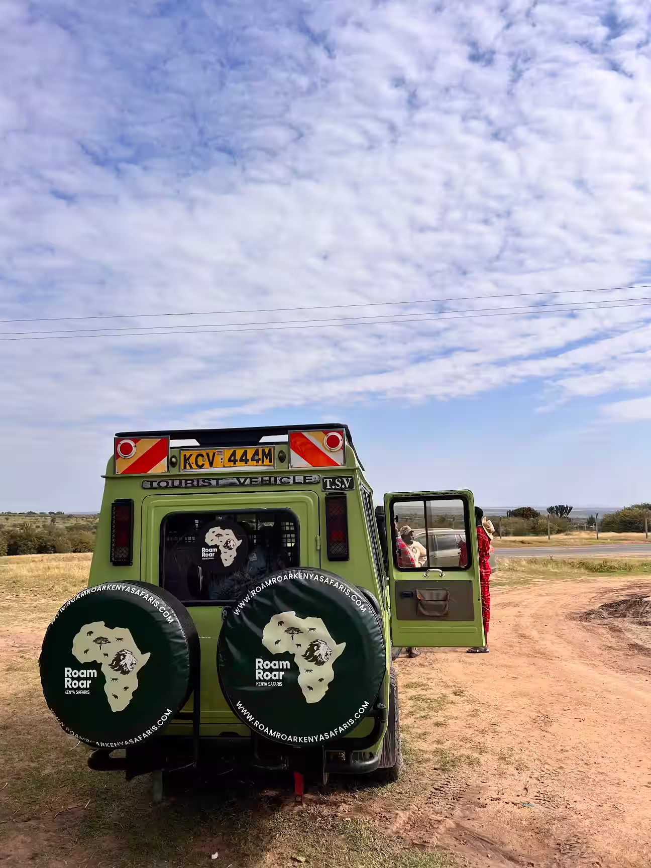 Rear view of a green safari vehicle with open door, two spare tires covered with Roam Roar Kenya Safaris logos, parked on a dirt road in a grassy savanna under a partly cloudy sky.