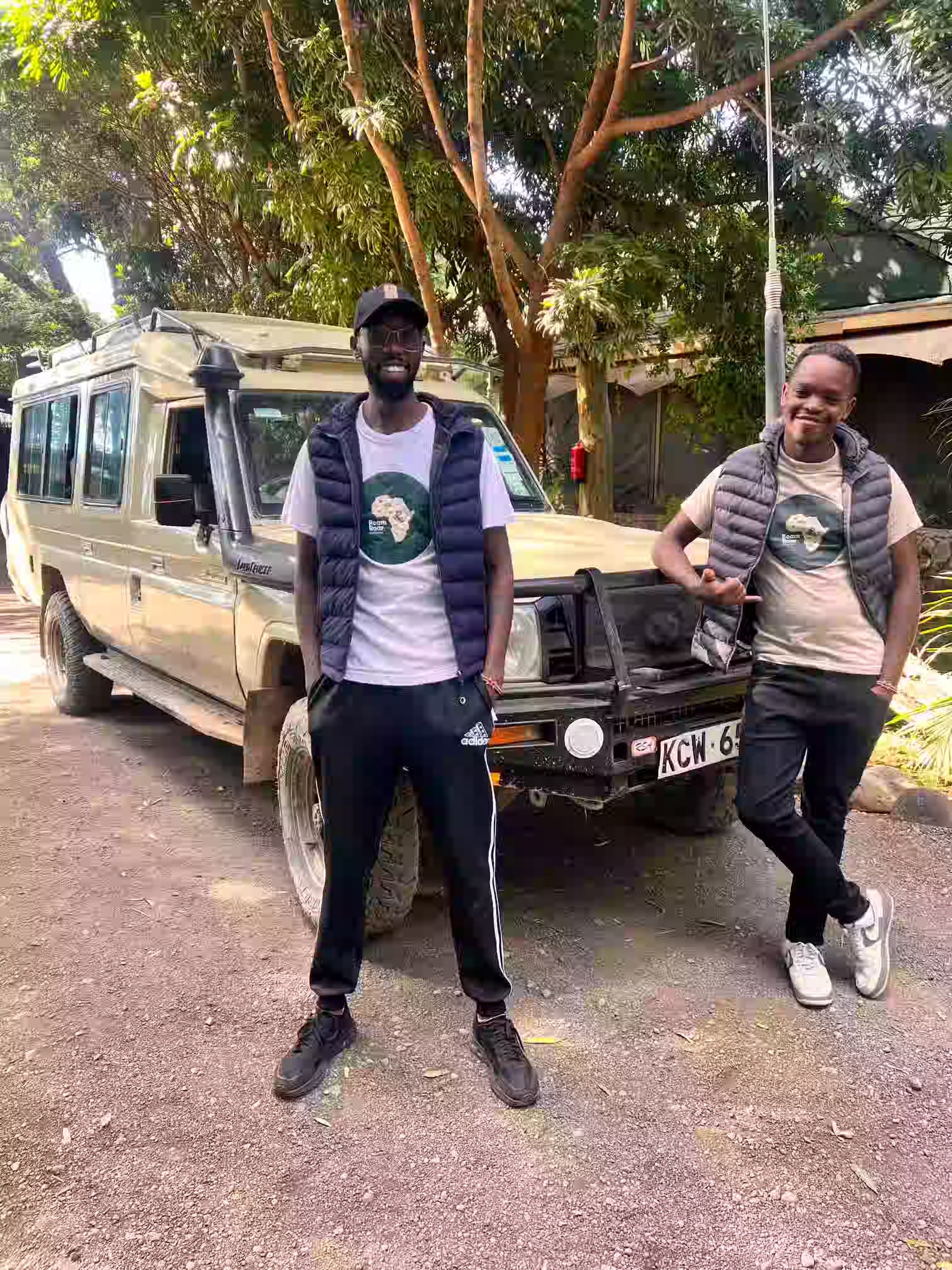 Two men smiling and posing in front of a beige safari vehicle under trees, both wearing puffer vests and t-shirts with an Africa logo.