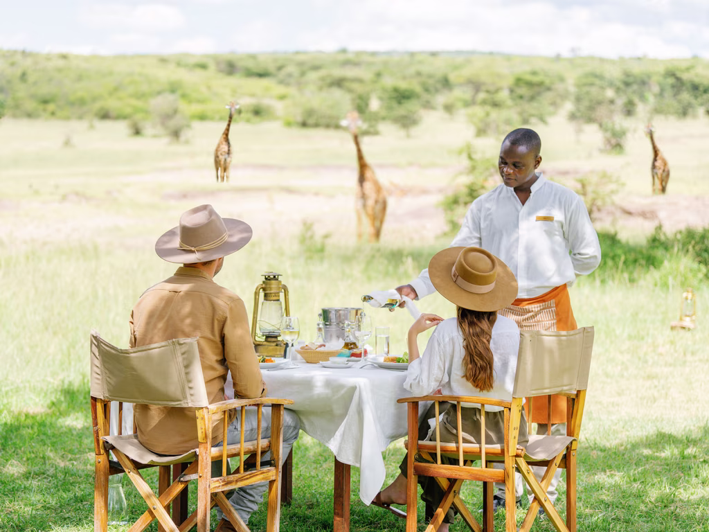 A couple wearing hats dining outdoors at a table with a white tablecloth while a waiter pours a drink, with giraffes visible in the grassy background.