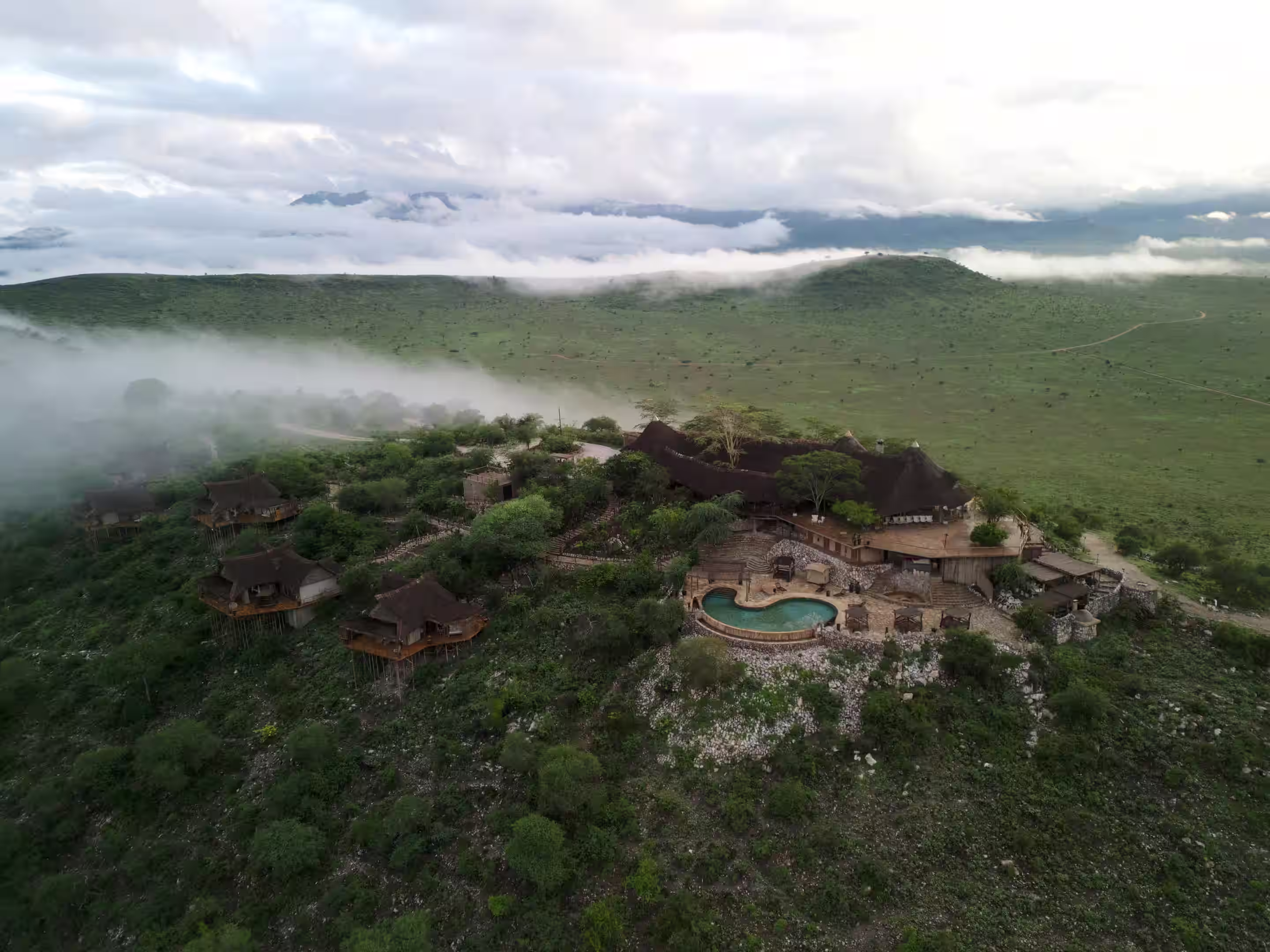 Aerial view of a hillside lodge with multiple thatched-roof cabins surrounded by greenery and mist in a vast savannah landscape.
