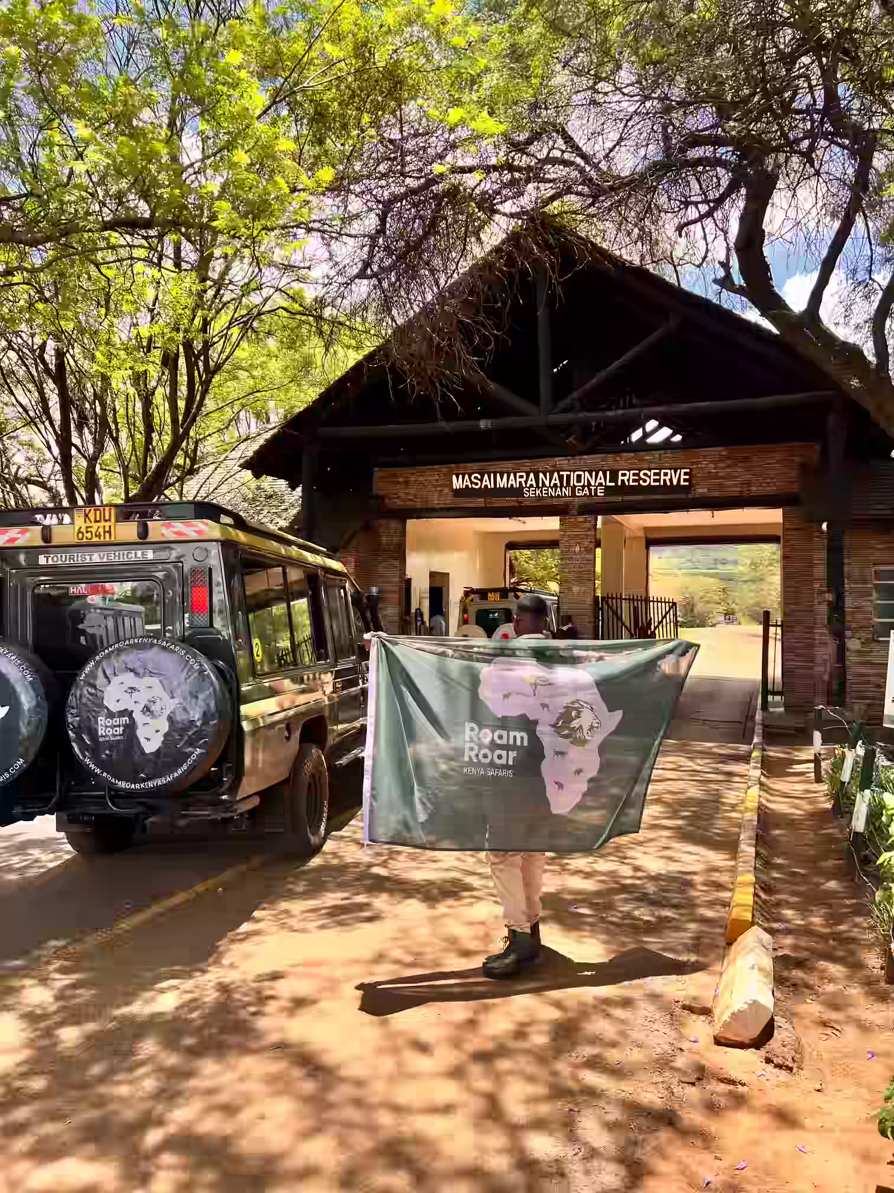 A guest holding a Roam Roar Kenya Safaris flag in front of Masai Mara National Reserve Sekenani Gate as RoamRoar safari vehicle enter the reserve