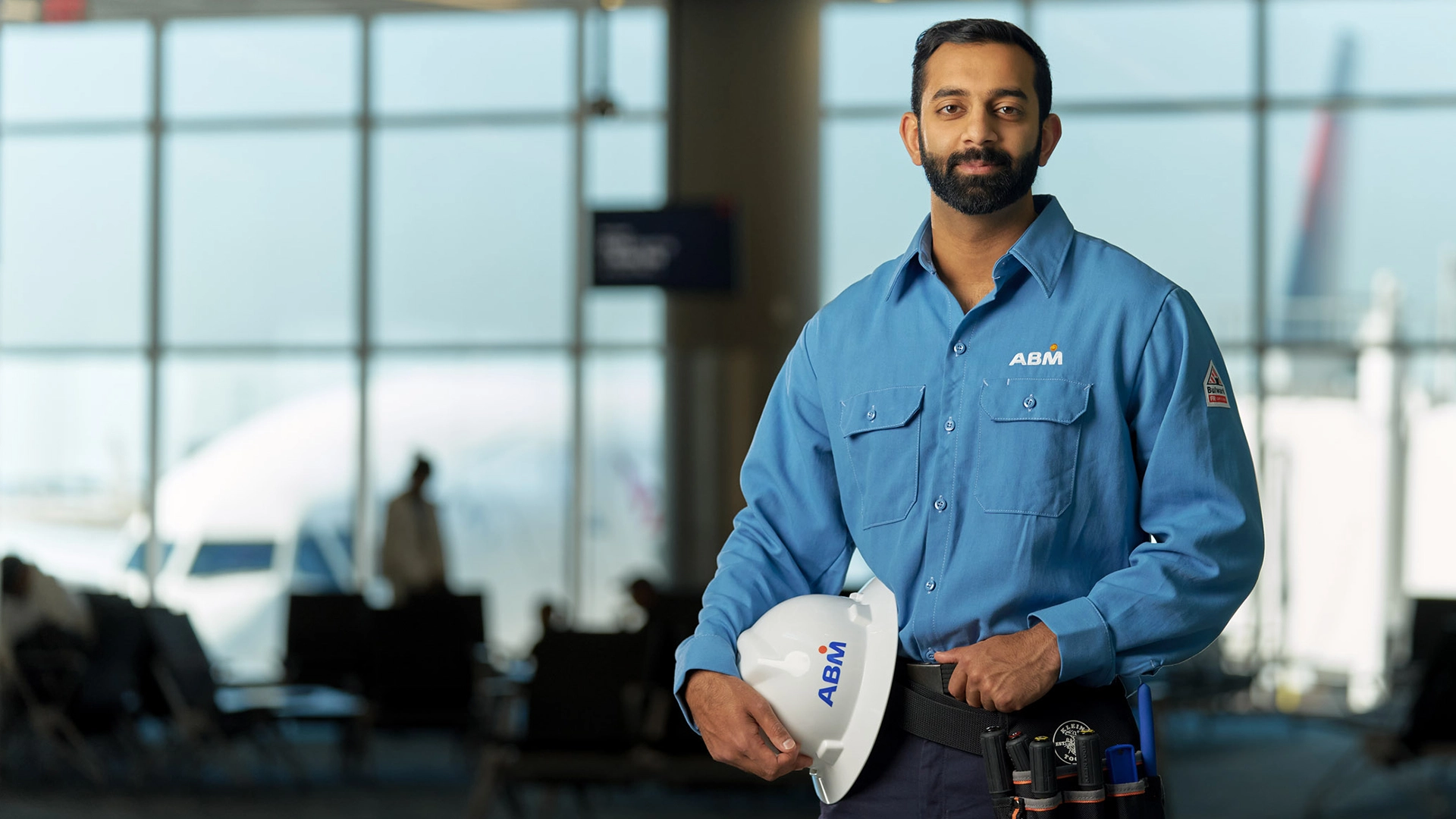 A man in an ABM uniform standing inside of an airport holding a hard hat by his side