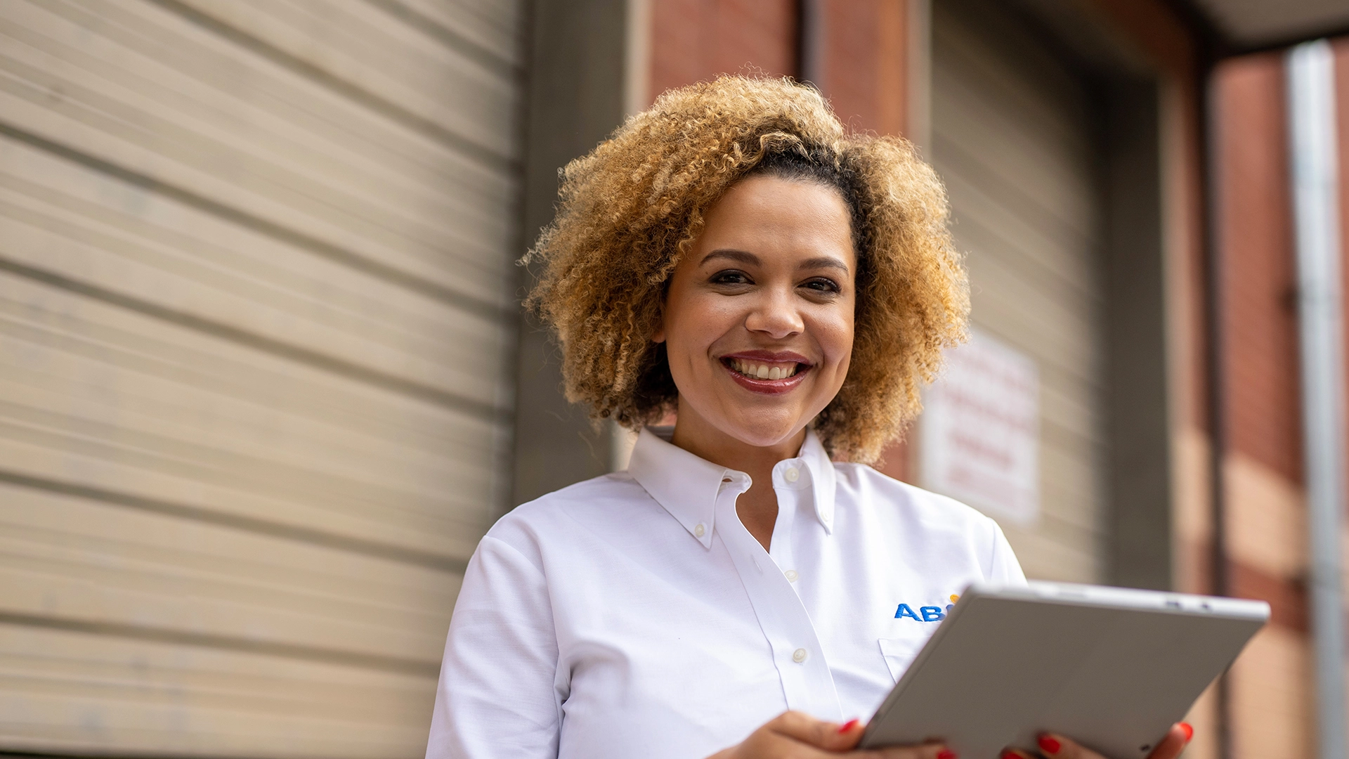 A woman smiling enthusiastically, holding a tablet in front of a warehouse loading dock