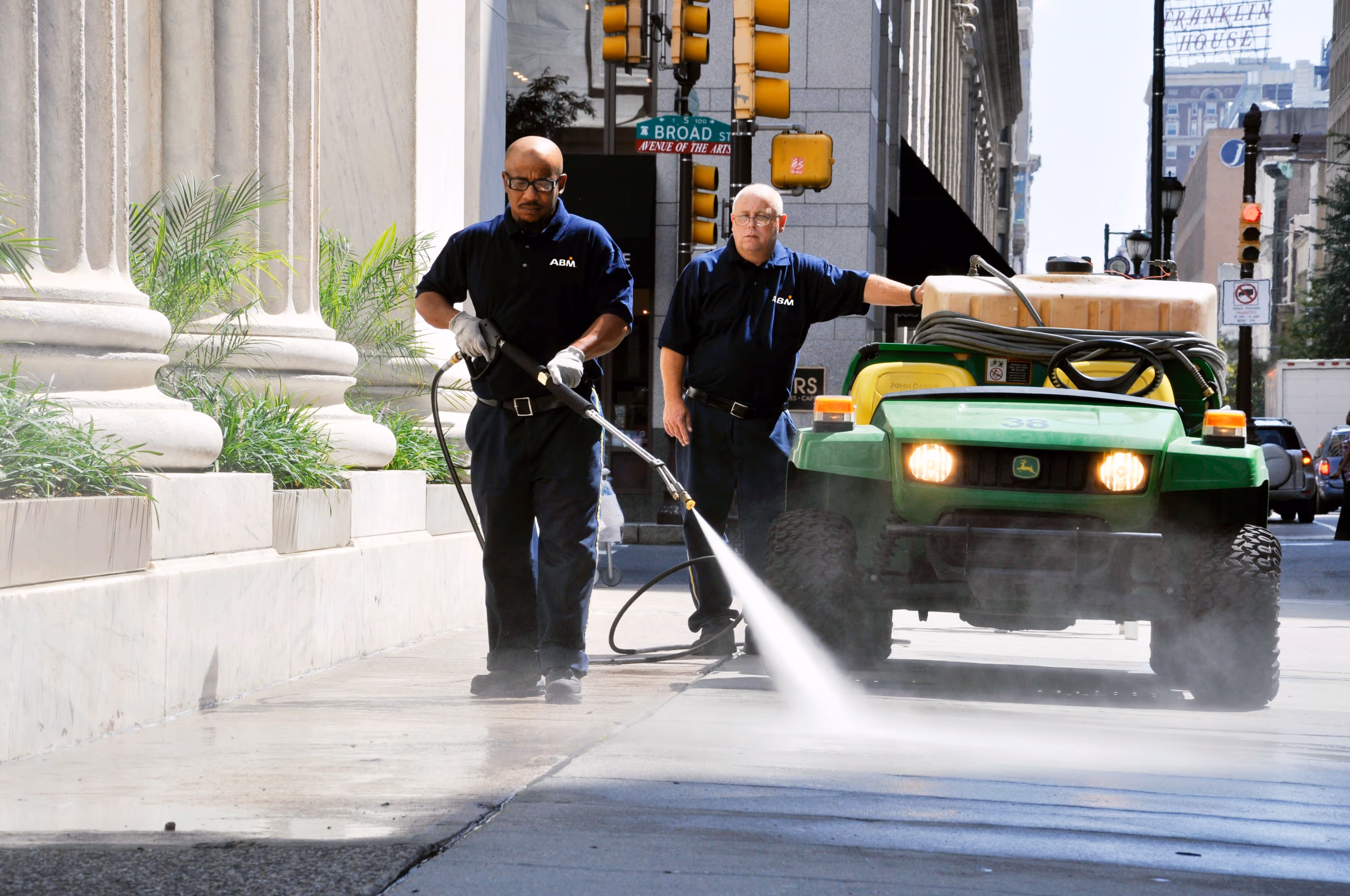 Two men outside, one by a John Deere vehicle, the other pressure washing the street
