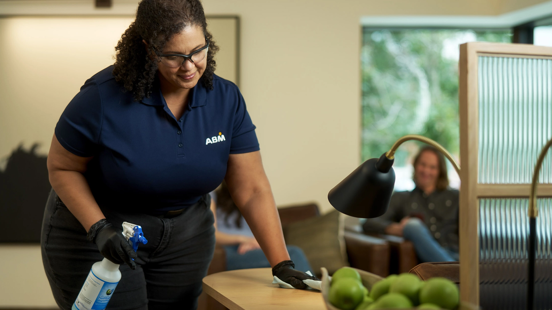 A woman in a collared ABM shirt holding a spray bottle and wiping a desk