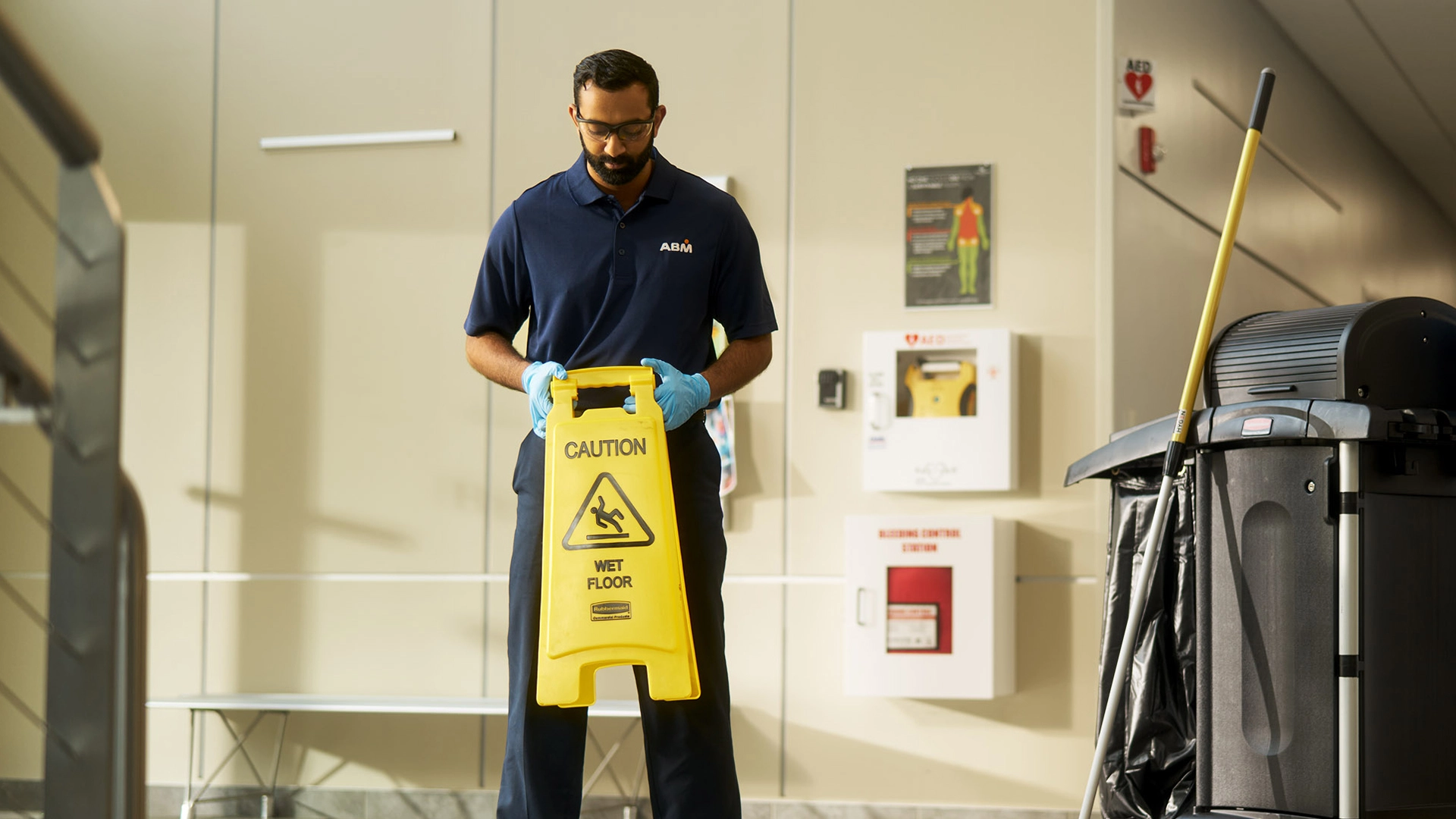 A person in an ABM uniform with gloves on, holding a caution sign next to cleaning equipment