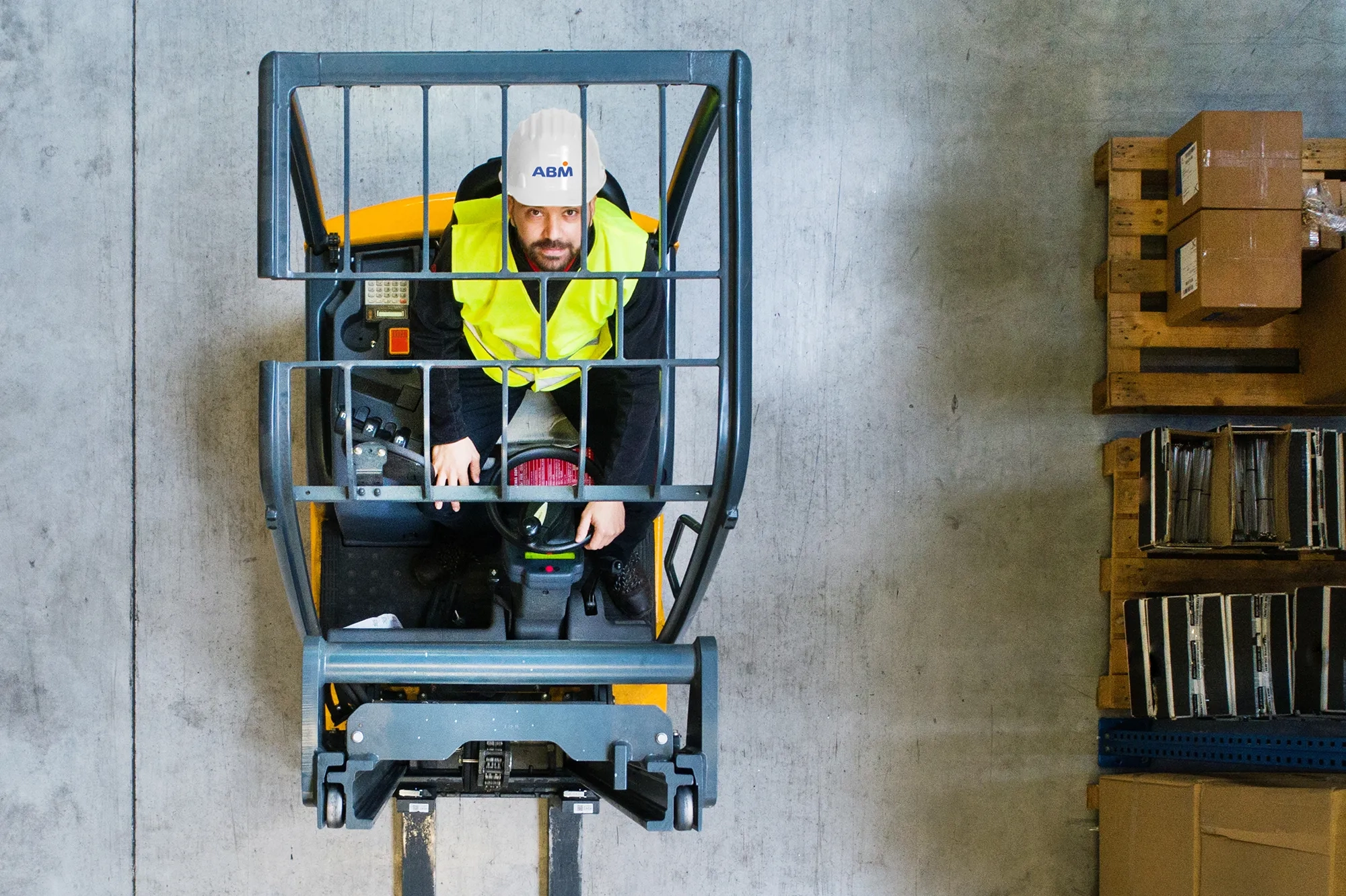 A person in an ABM hardhat and reflective vest driving a forklift, looking straight up towards the camera