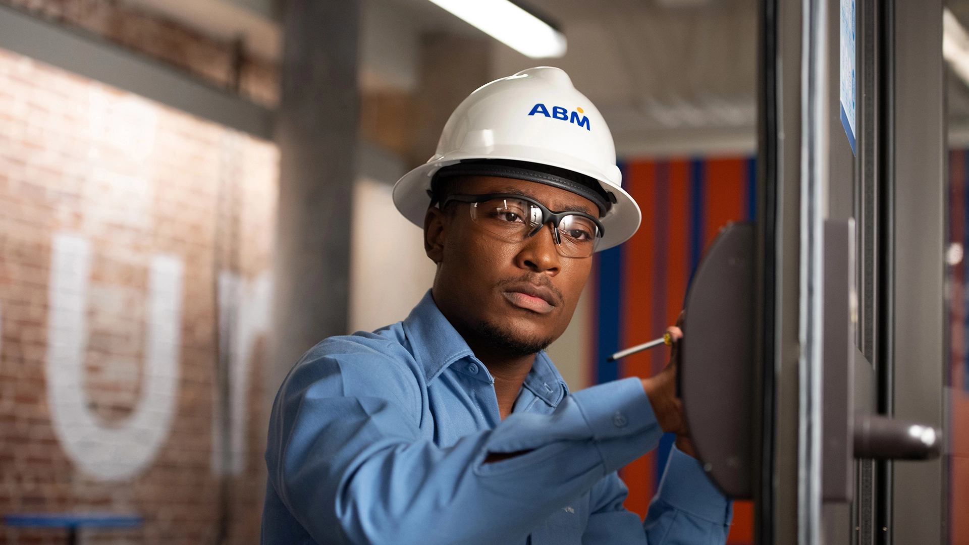 ABM worker wearing a hard hat and safety glasses adjusting a wall-mounted device inside an industrial building