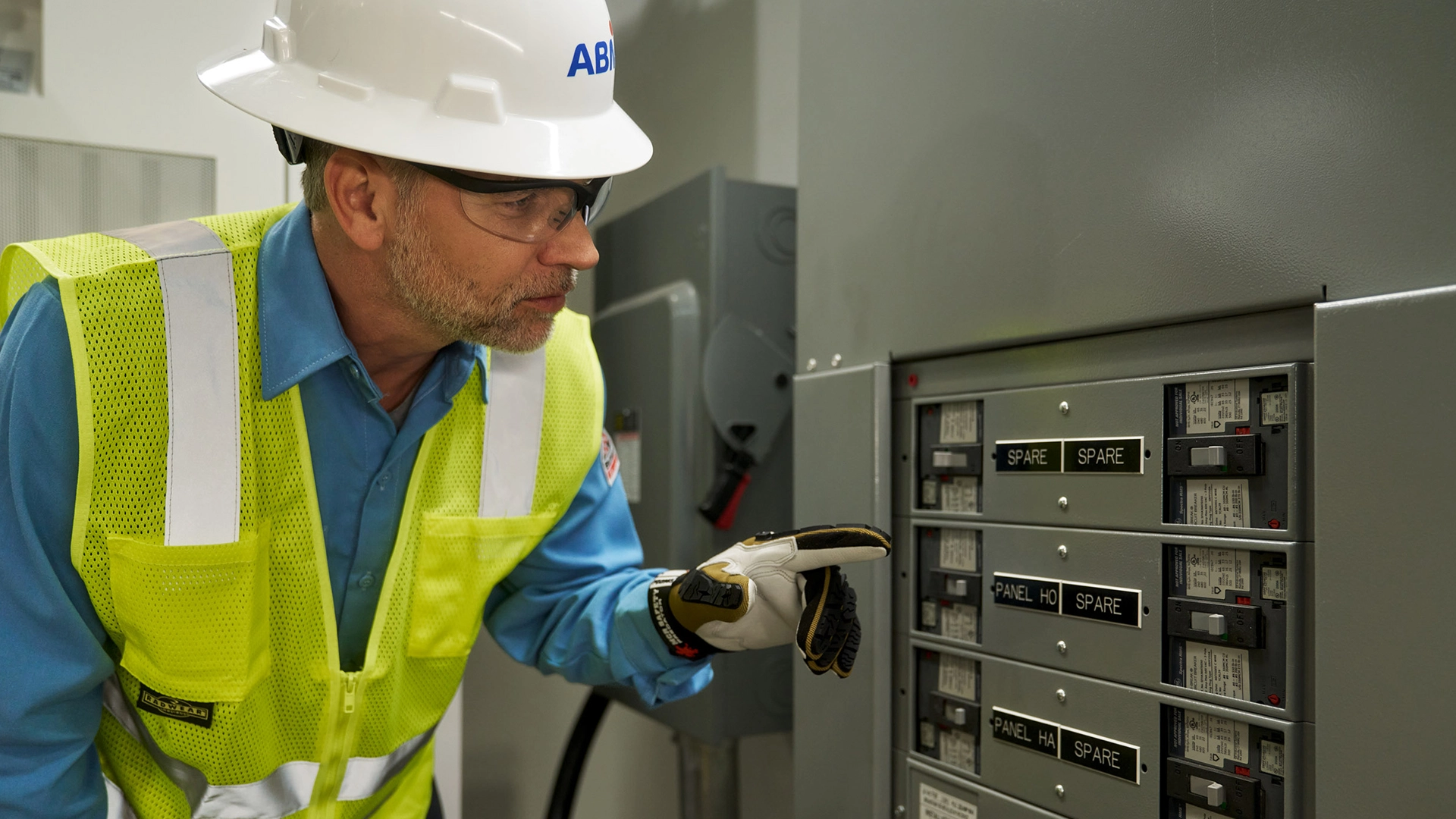 ABM maintenance worker wearing a hard hat and safety vest inspects an electrical panel