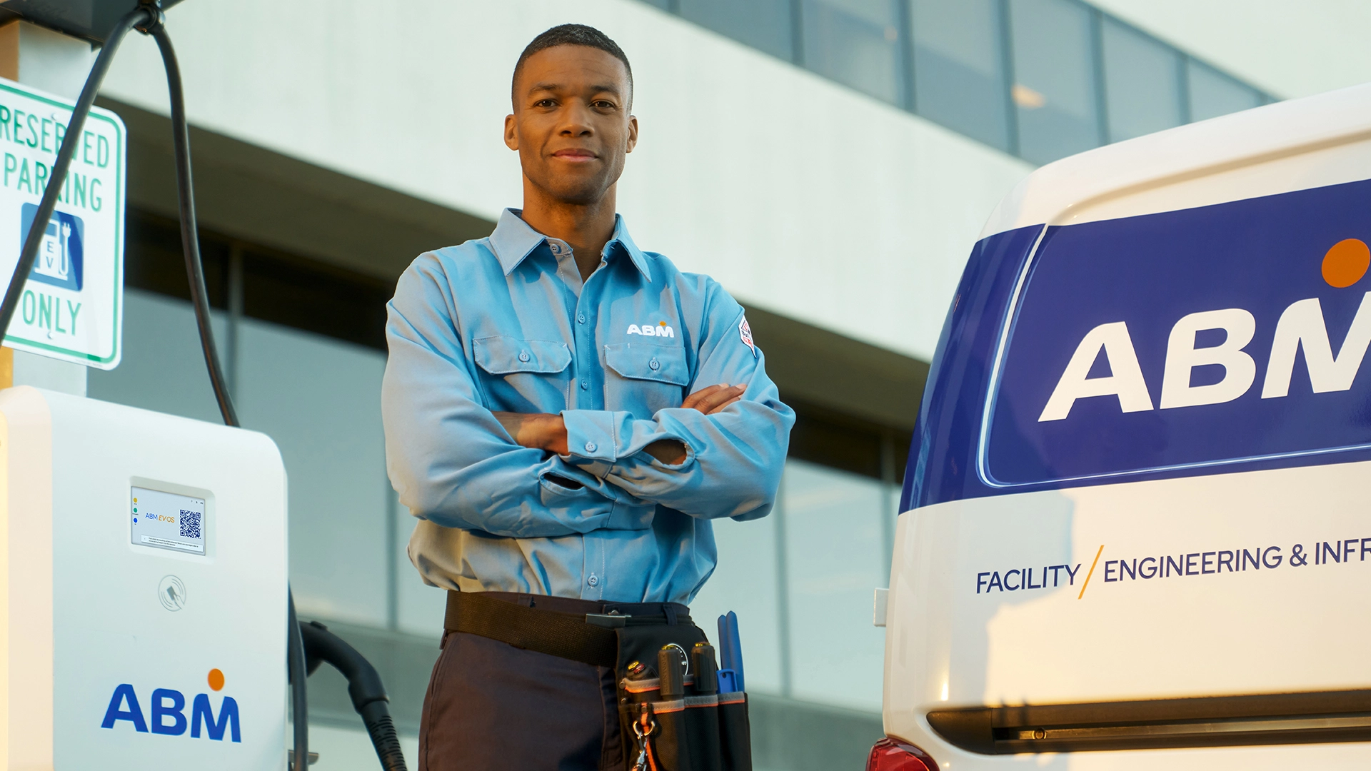 An ABM technician in a blue uniform stands with arms crossed beside a branded service van and an electric vehicle charging station