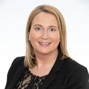 A smiling woman with shoulder-length blonde hair, wearing a black blazer over a lace top, against a light background.