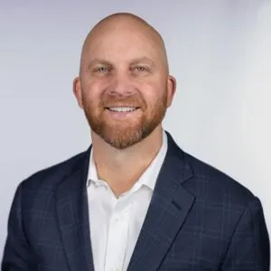 A smiling man with a bald head and a beard, wearing a dark blazer over a white shirt, against a light background.