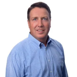 A man with short hair and blue eyes smiles, wearing a light blue checkered shirt against a white background.