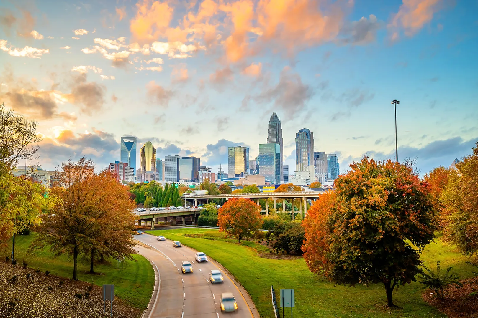 Charlotte city skyline at sunset with autumn-colored trees and cars on the highway.