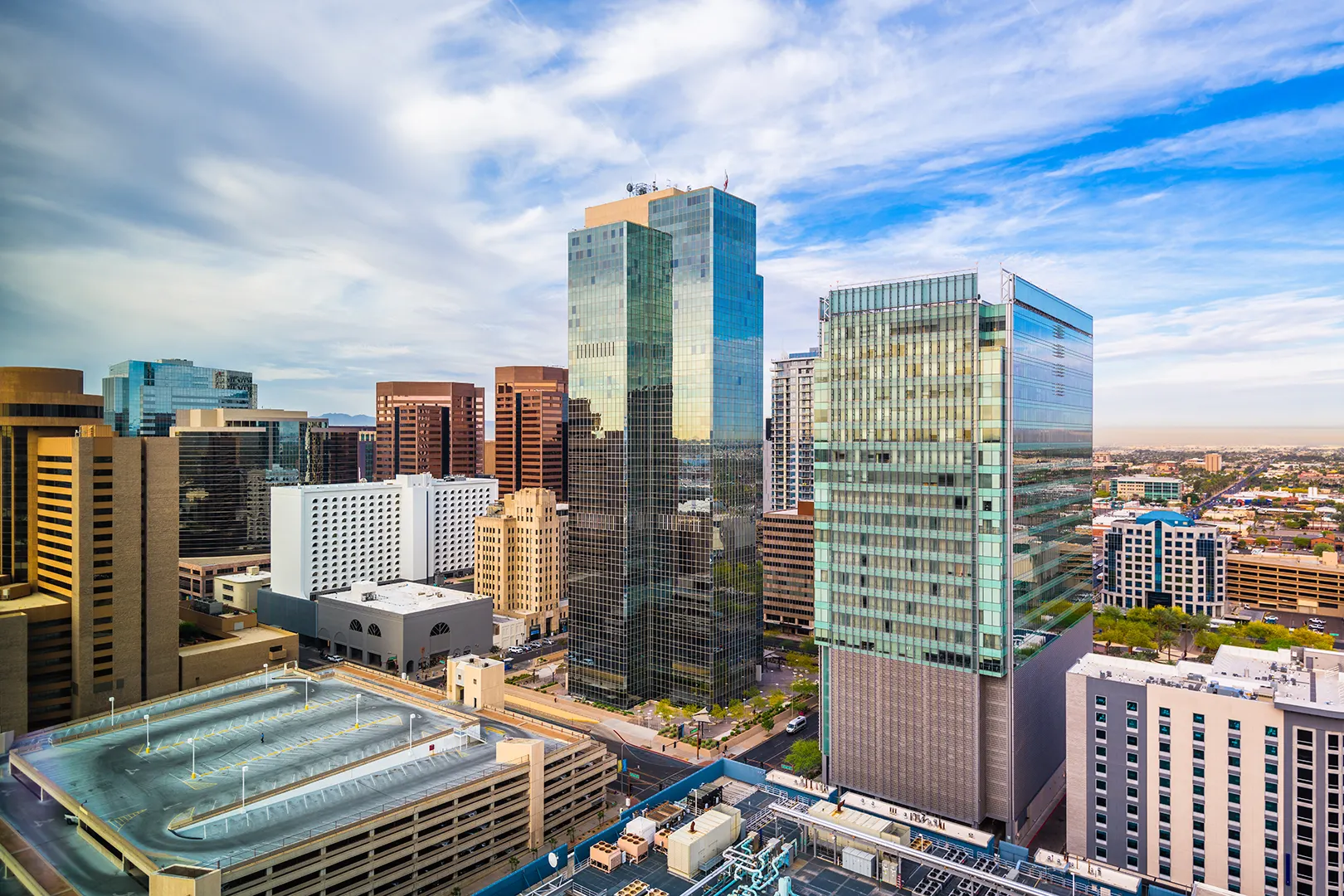Downtown cityscape with modern glass skyscrapers and a multi-level parking garage under a partly cloudy blue sky.