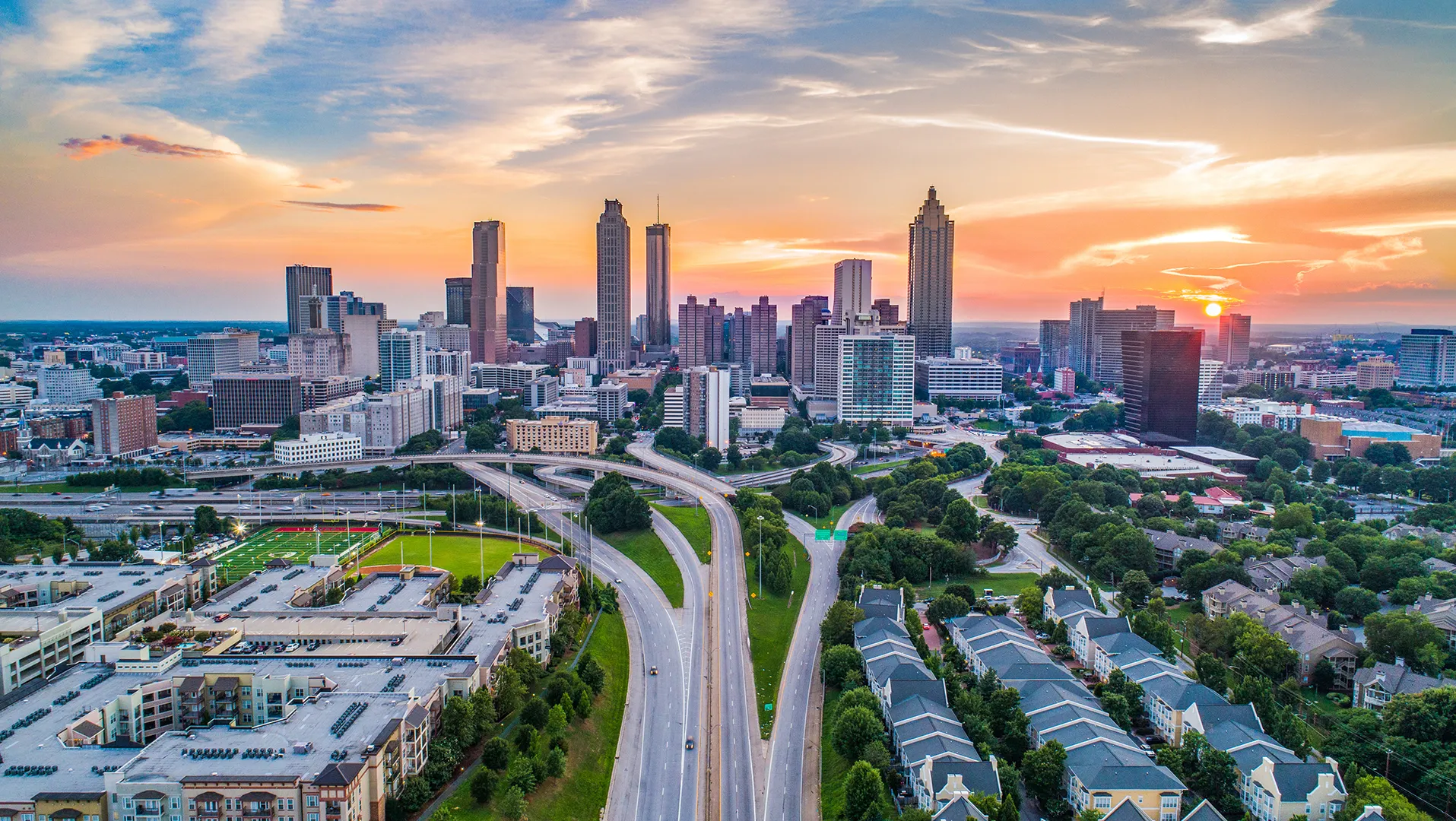 Aerial view of a city skyline with highways, residential buildings, and a sunset in the background.