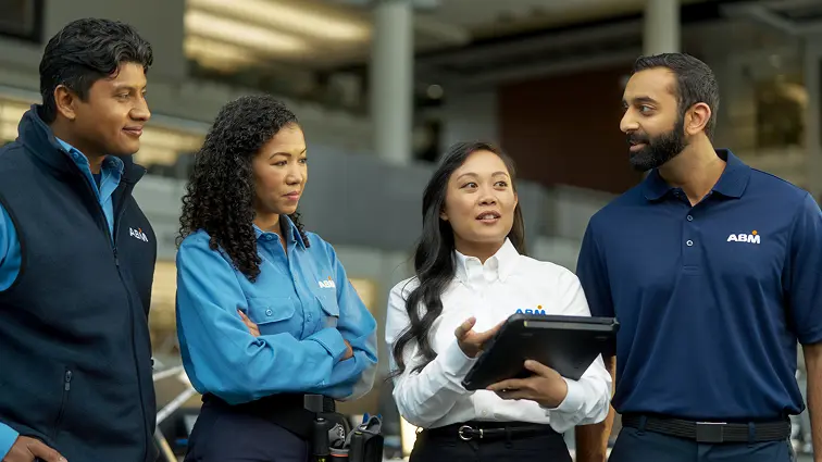 Four ABM employees in branded uniforms collaborating indoors, with one holding a tablet and speaking to the group.