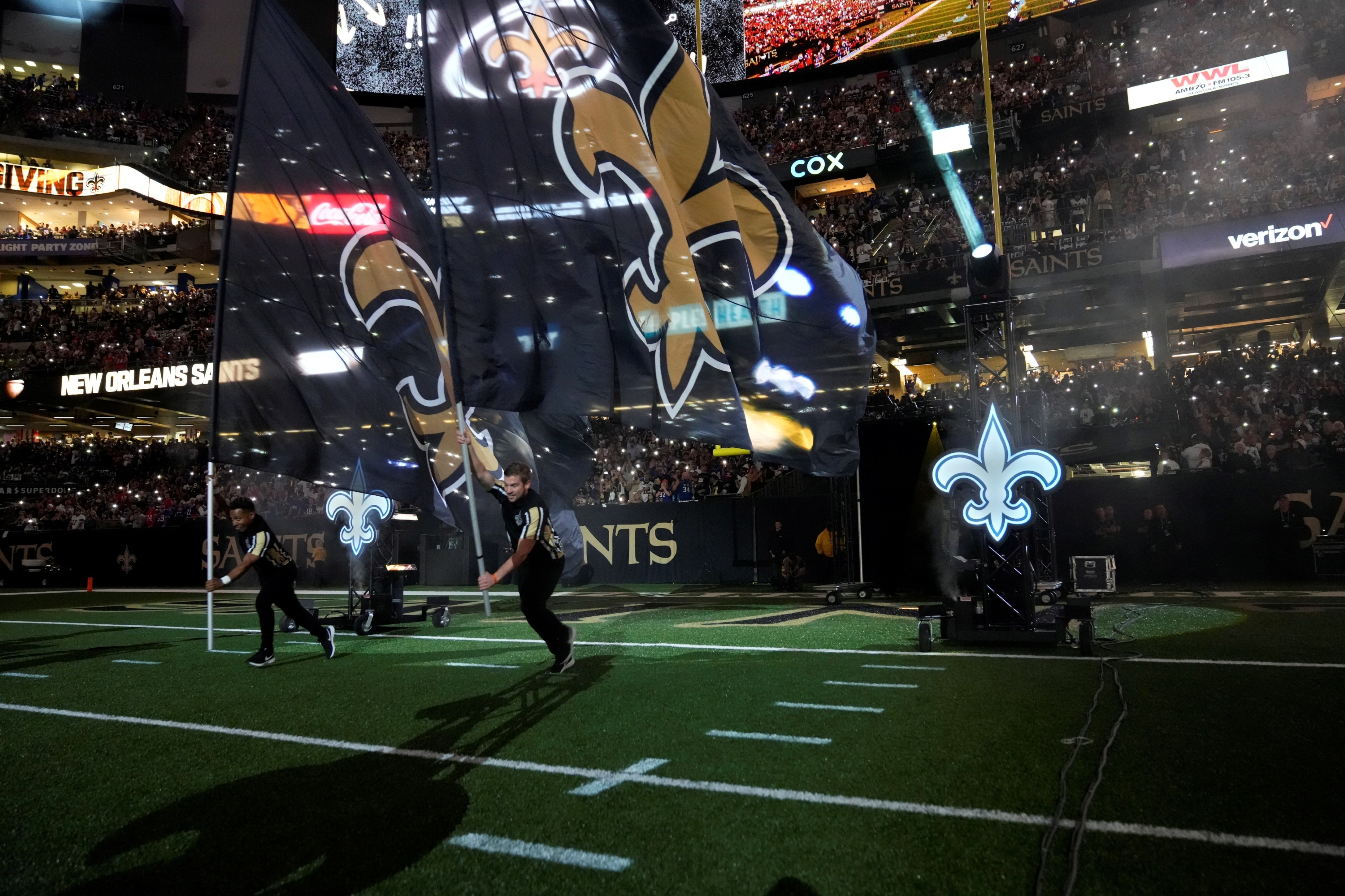 Two flag bearers run across the football field carrying large black flags with the New Orleans Saints' gold fleur-de-lis logo. The stadium is packed with cheering fans, illuminated lights, and team branding displays during a game-day introduction.