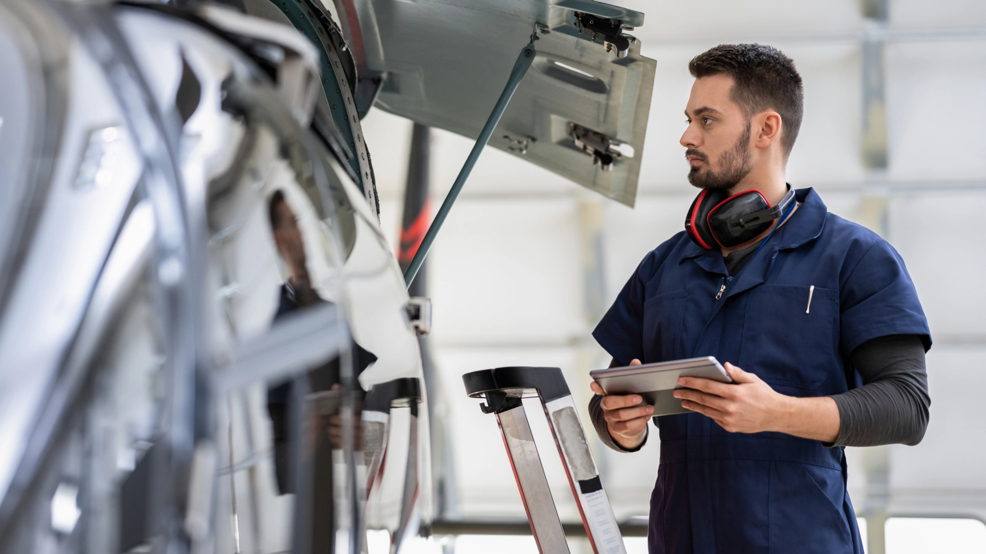 Technician in navy coveralls holding a tablet and inspecting an aircraft in a maintenance hangar.