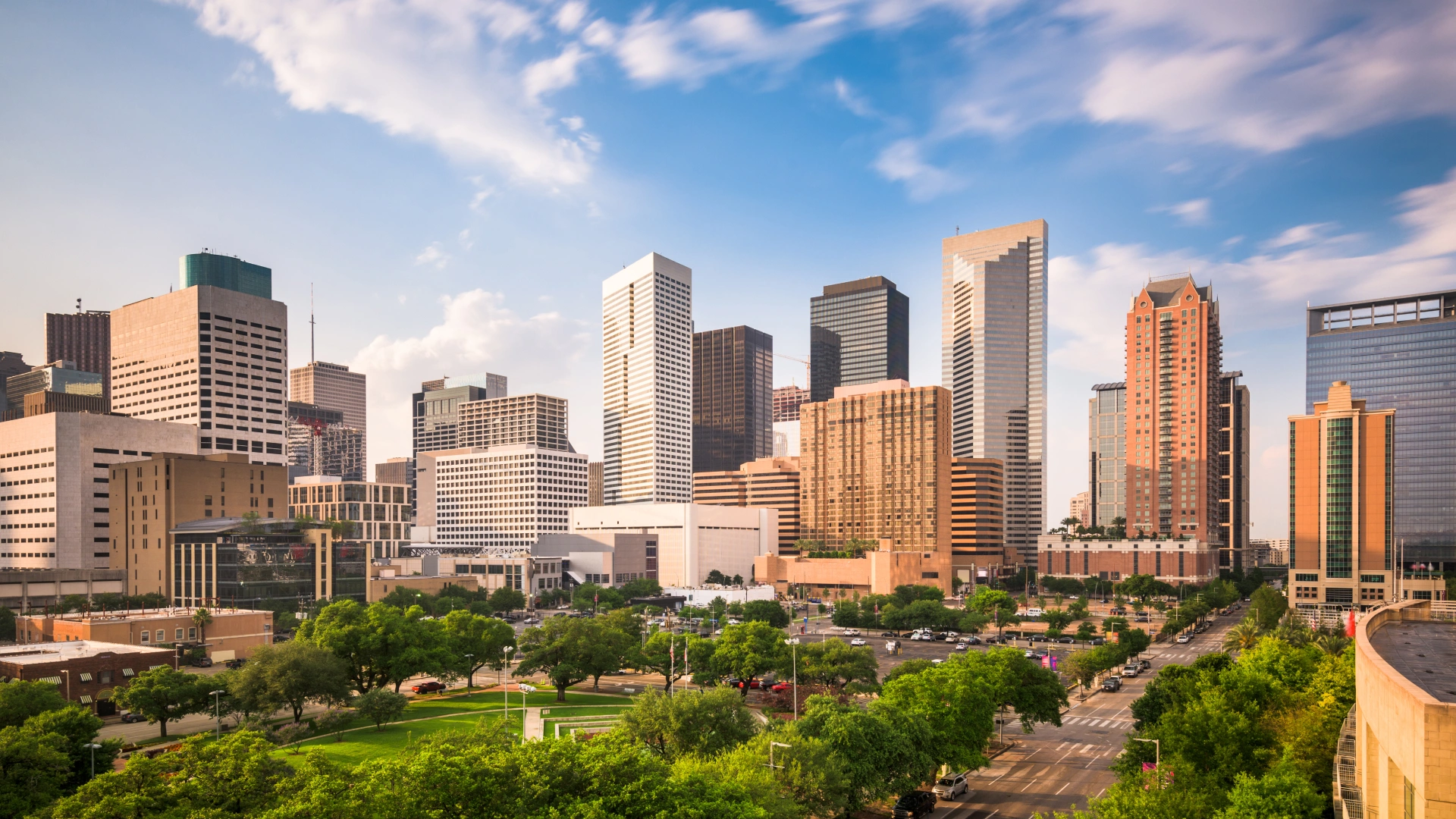 City skyline with various modern office buildings under a partly cloudy blue sky and a green park in the foreground.