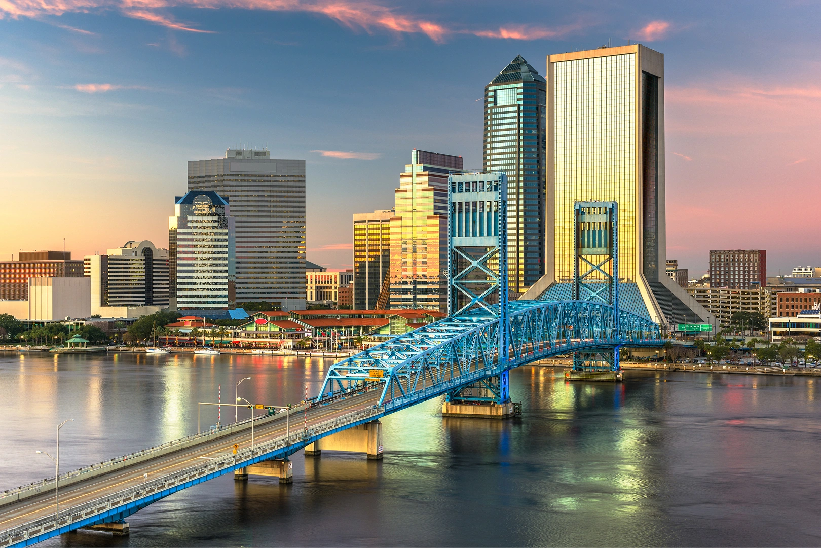 Blue steel bridge crossing a calm river towards a city skyline with tall buildings reflecting sunset light.