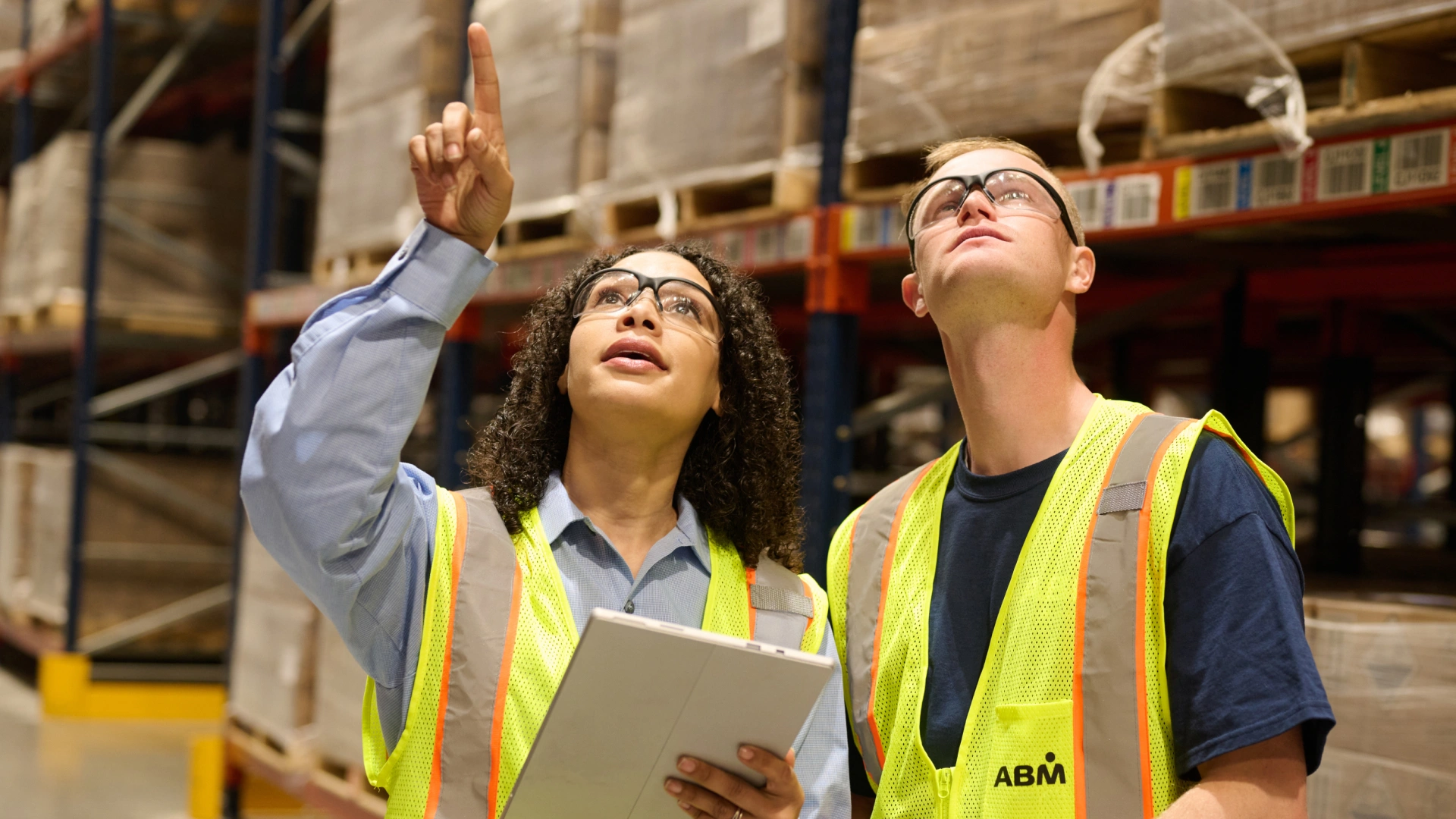 Two warehouse workers wearing safety glasses and yellow safety vests inspecting inventory with one pointing upward and the other looking up.