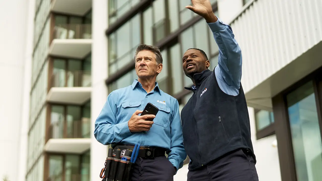 Two ABM workers, one in a white shirt holding a tablet pointing upwards and the other in a blue uniform and hard hat listening attentively in a building corridor.