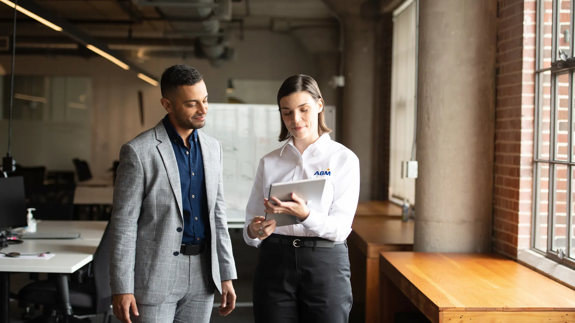 Two professionals in an office, one in a white ABM shirt showing a tablet to a man in a gray suit.