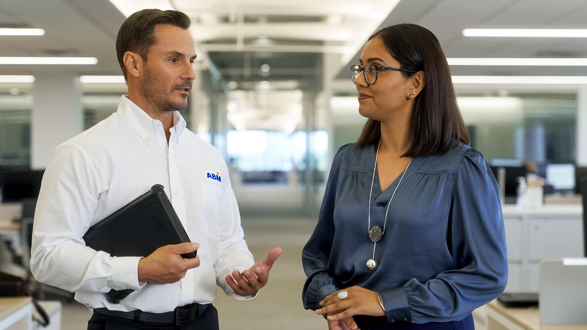 Man in ABM shirt holding tablet talking to woman in glasses and blue blouse in modern office.