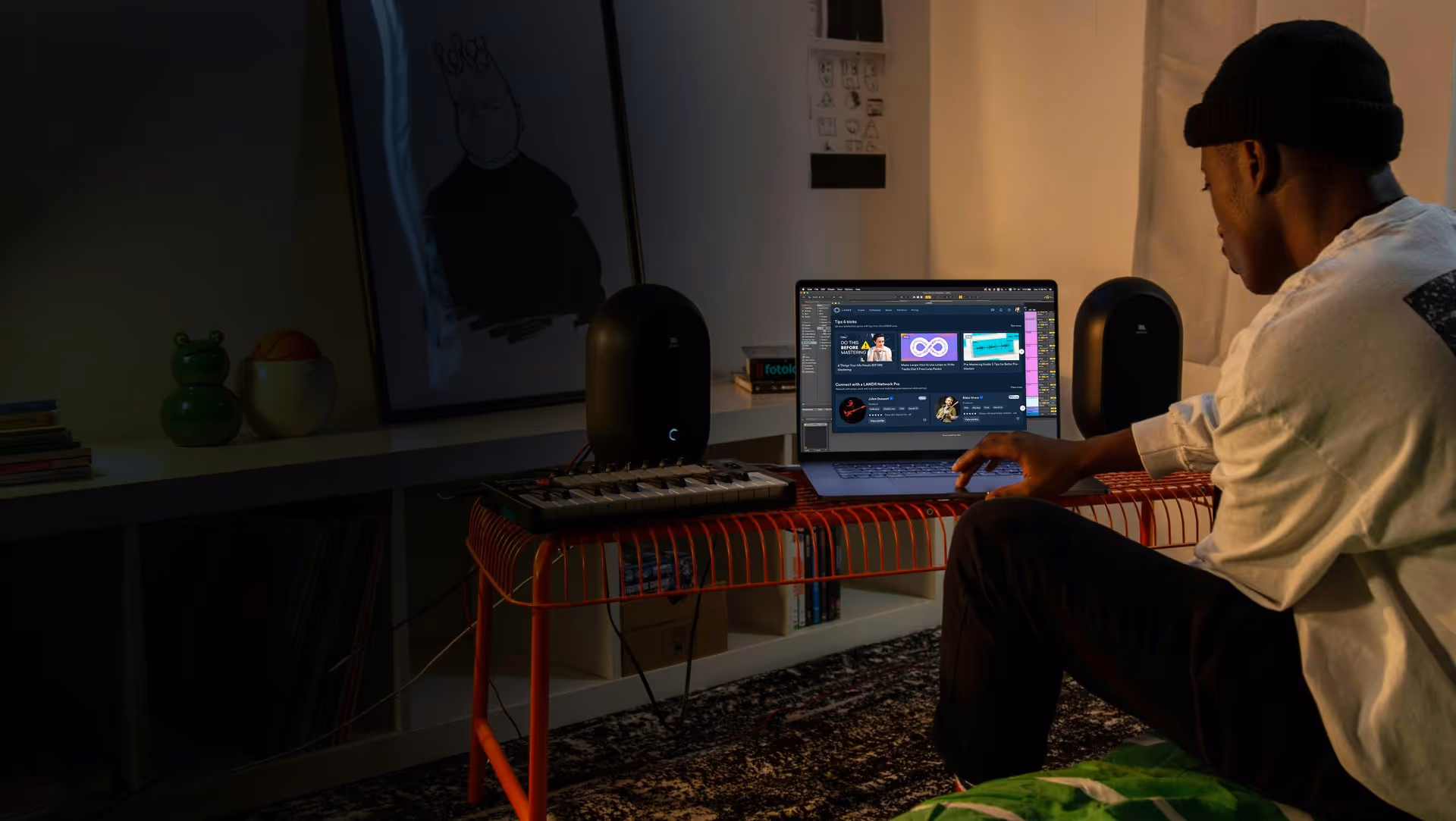 Person wearing a beanie using a laptop on a red wire table with two black speakers and a MIDI keyboard in a dimly lit room.