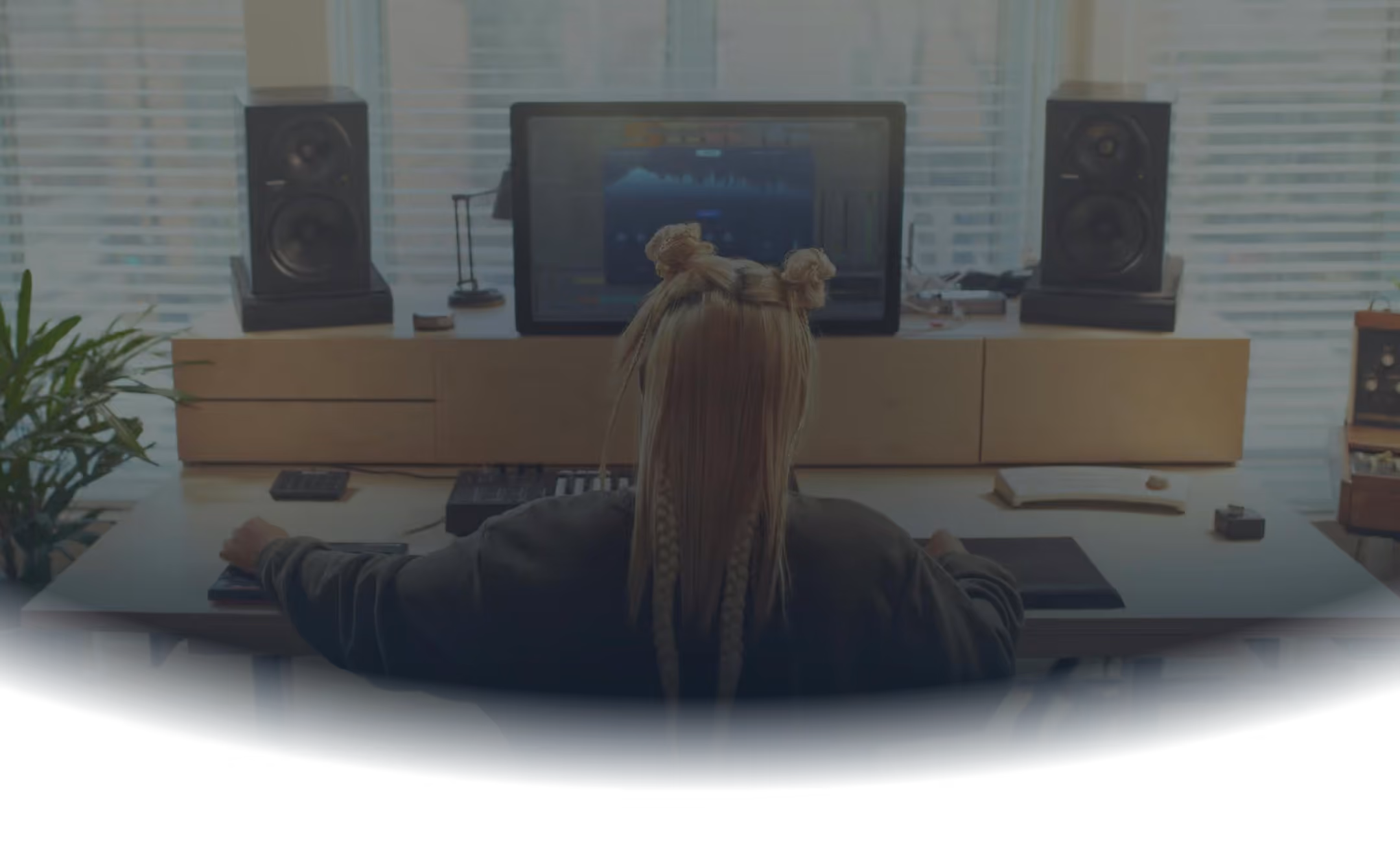 Person with braided hair styled in two buns working at a music production setup with speakers and a computer screen displaying audio software.