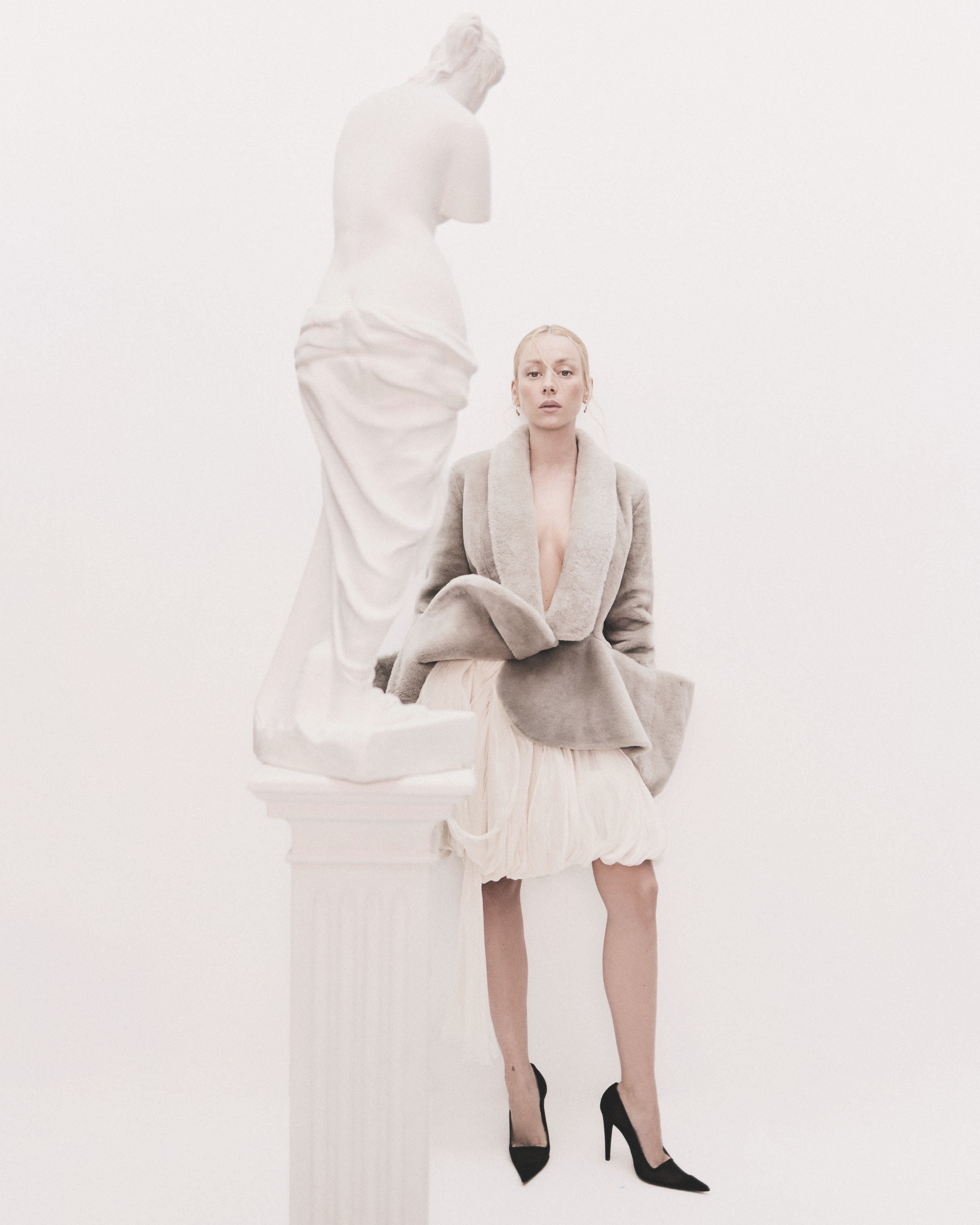 Ester Expósito in a pale shearling coat and black pumps beside a classical sculpture on a white set.