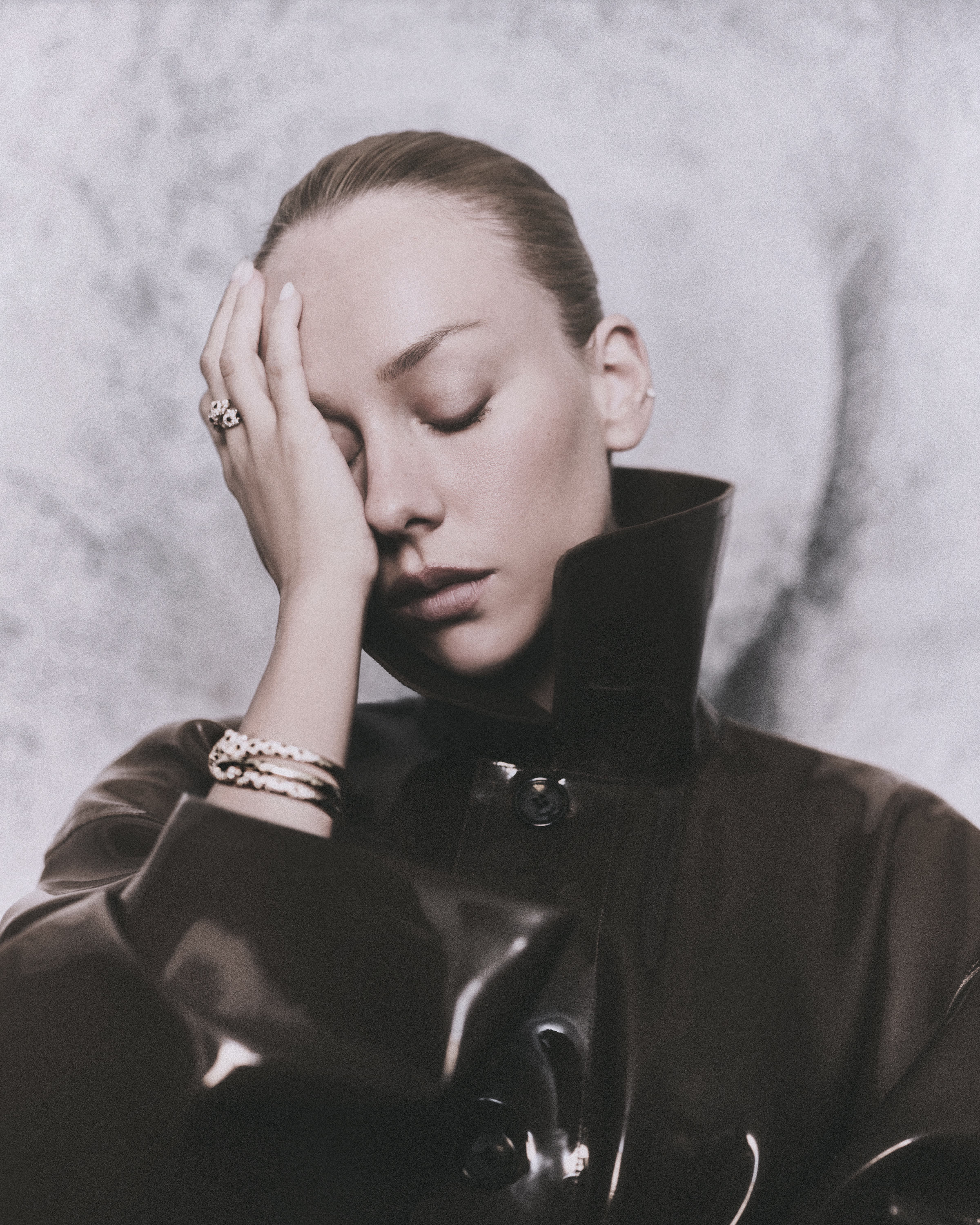 Close portrait of Ester Expósito in a glossy dark high-collared coat, wearing a ring and layered bracelets.
