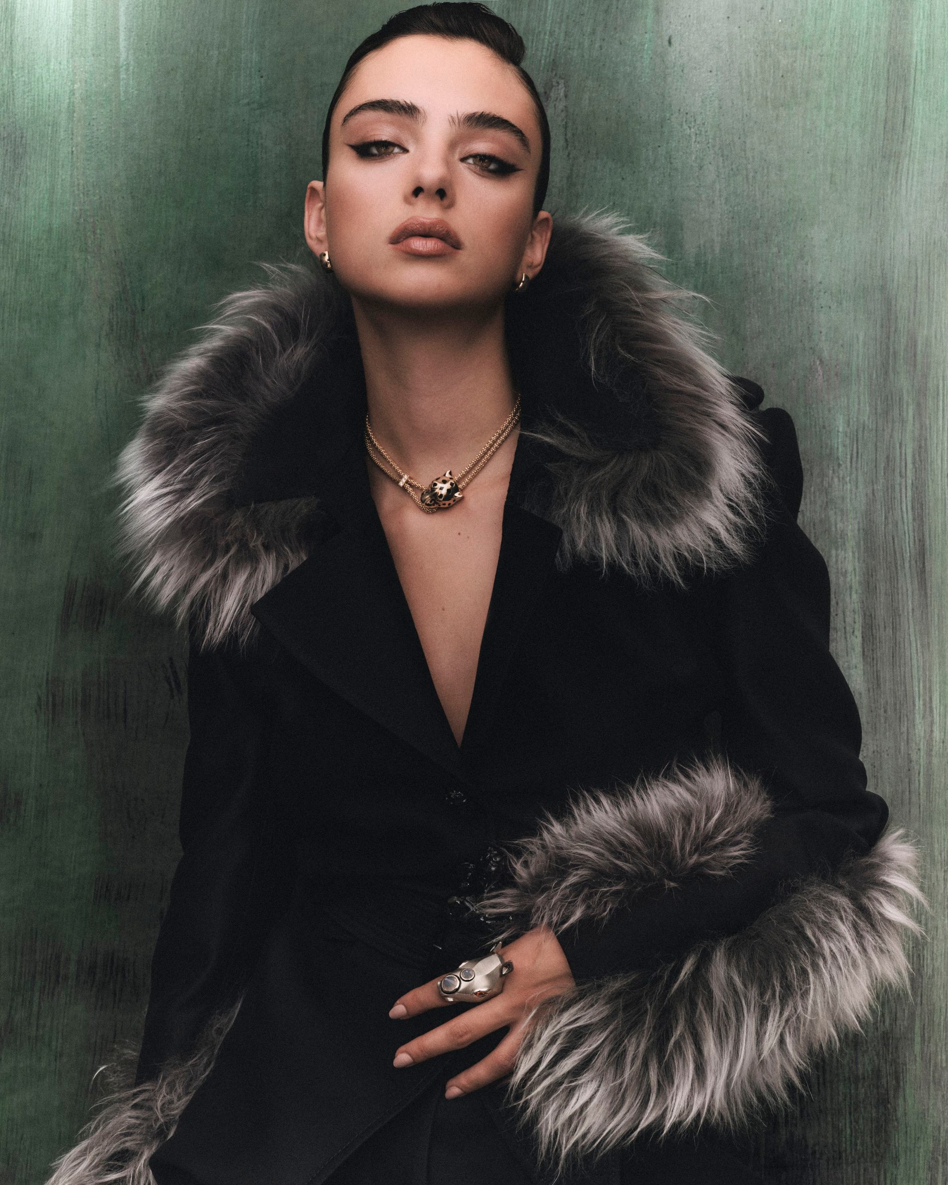 Deva Cassel in a dark fur-trimmed look against a muted green backdrop.