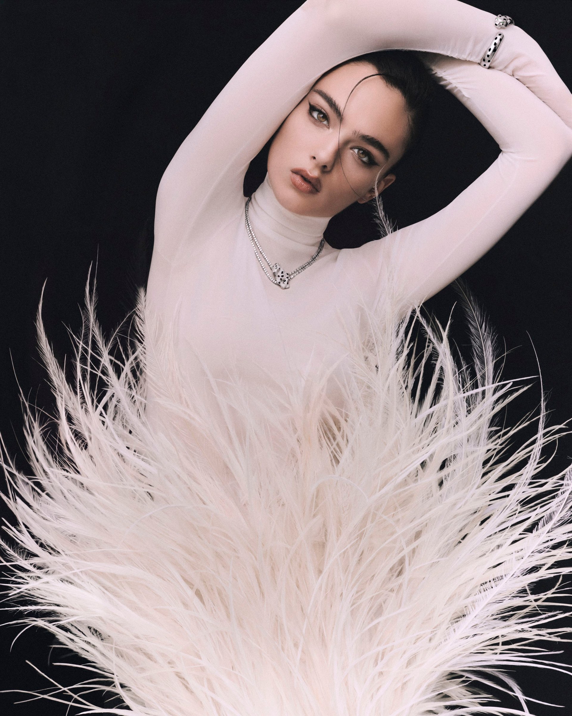 Deva Cassel in a pale high-neck look with both arms raised above white feathers.