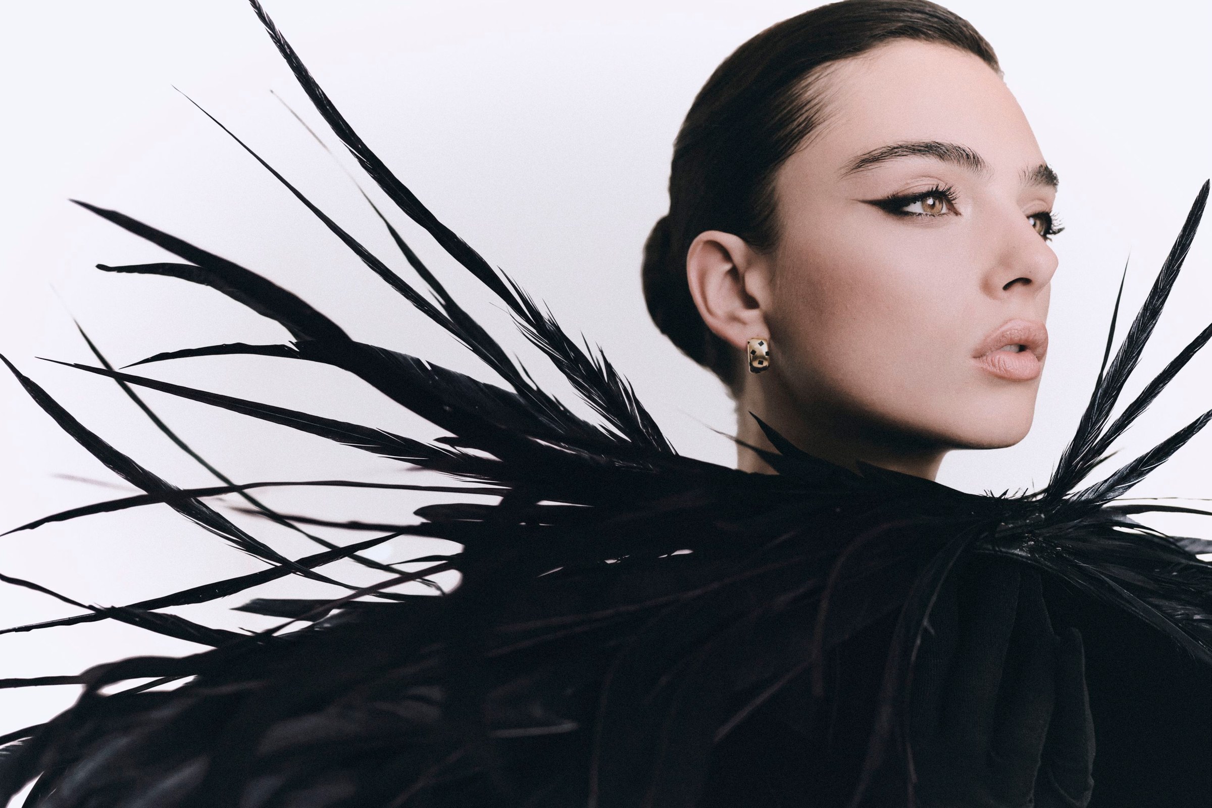 Wide profile portrait of Deva Cassel surrounded by black feathers against a pale backdrop.