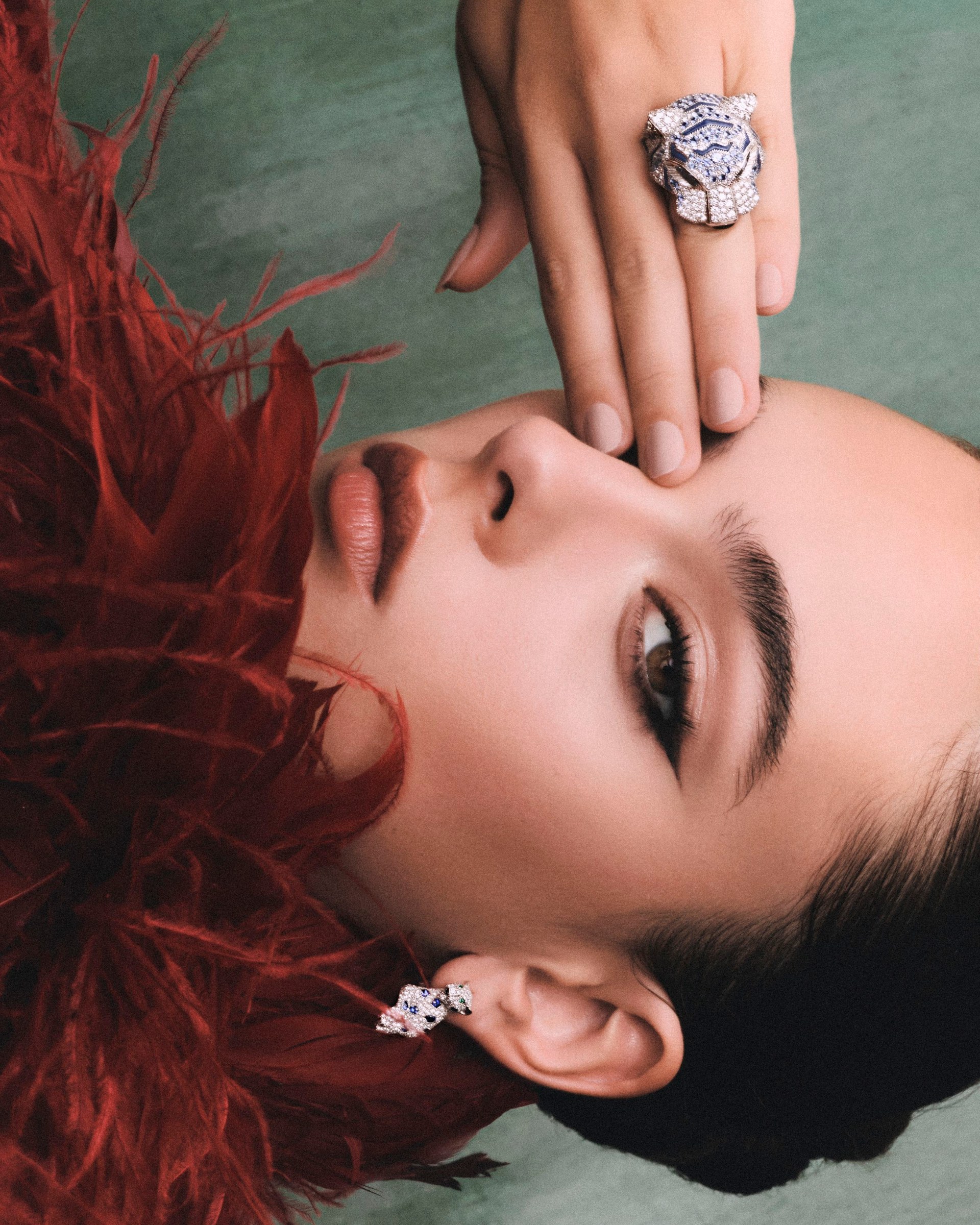 Sideways portrait of Deva Cassel among red feathers, a jewelled hand resting over one eye.