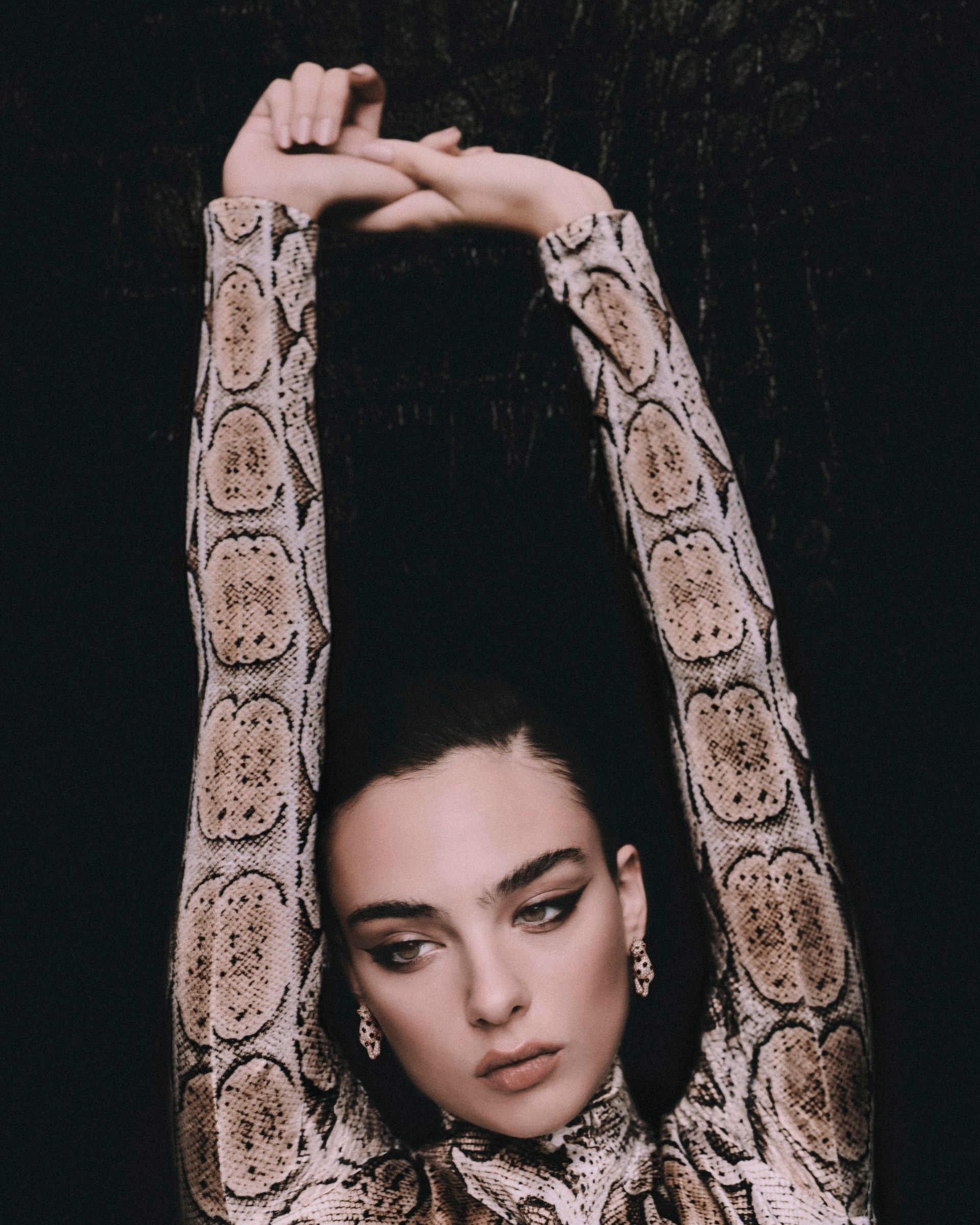 Deva Cassel in a snakeskin-patterned look with her arms raised against a dark backdrop.